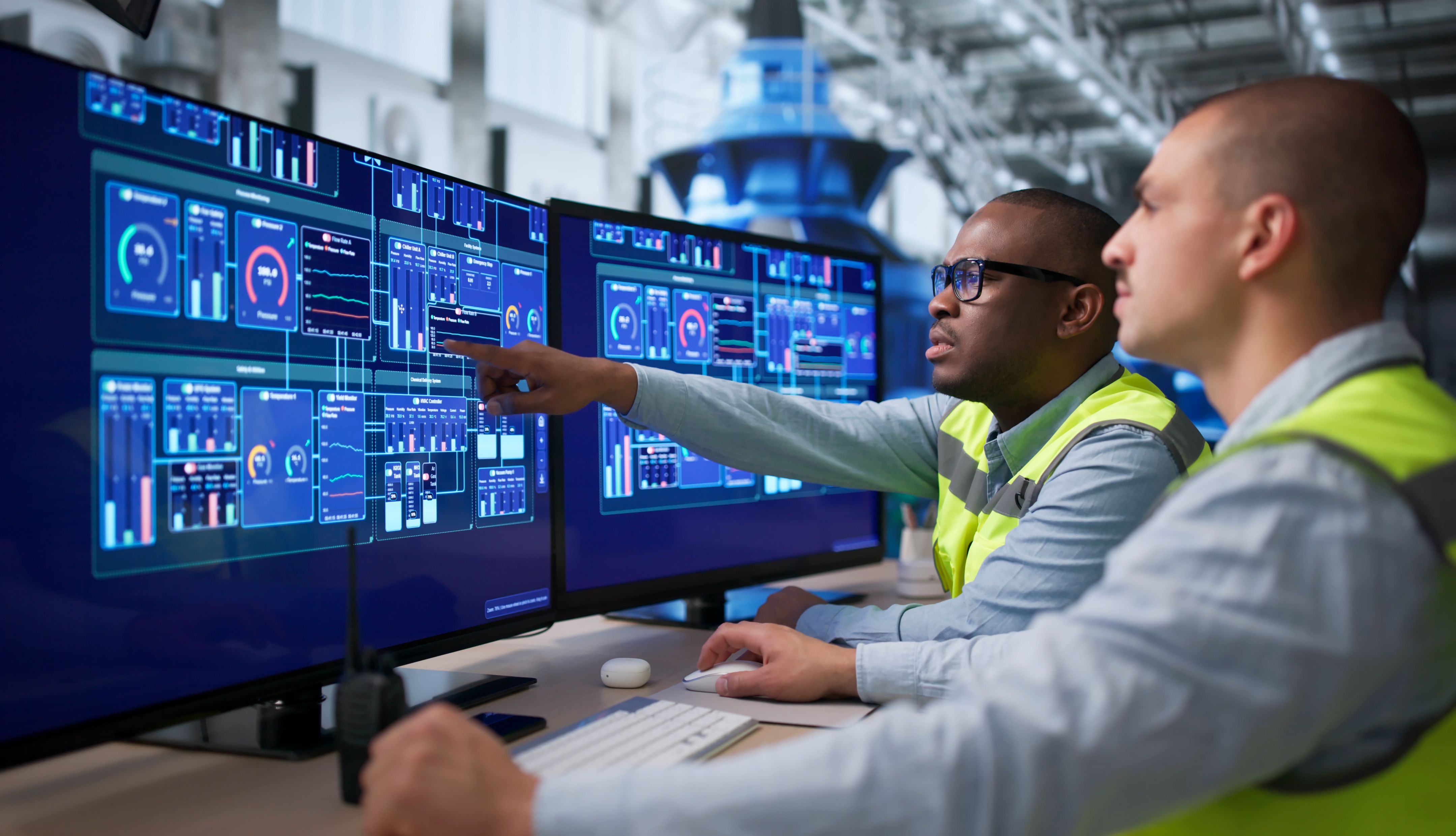Two workers in safety vests monitor digital manufacturing systems on multiple computer screens in an industrial facility. 