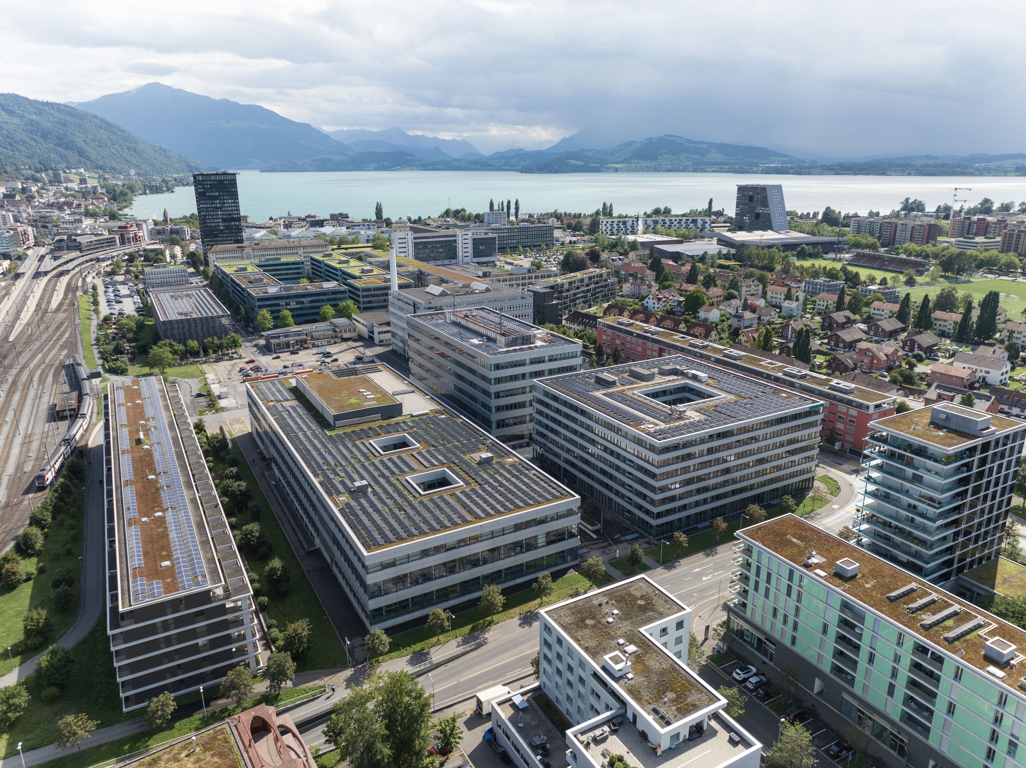 Aerial view of modern office buildings beside a lake, with mountains and cloudy skies in the background.