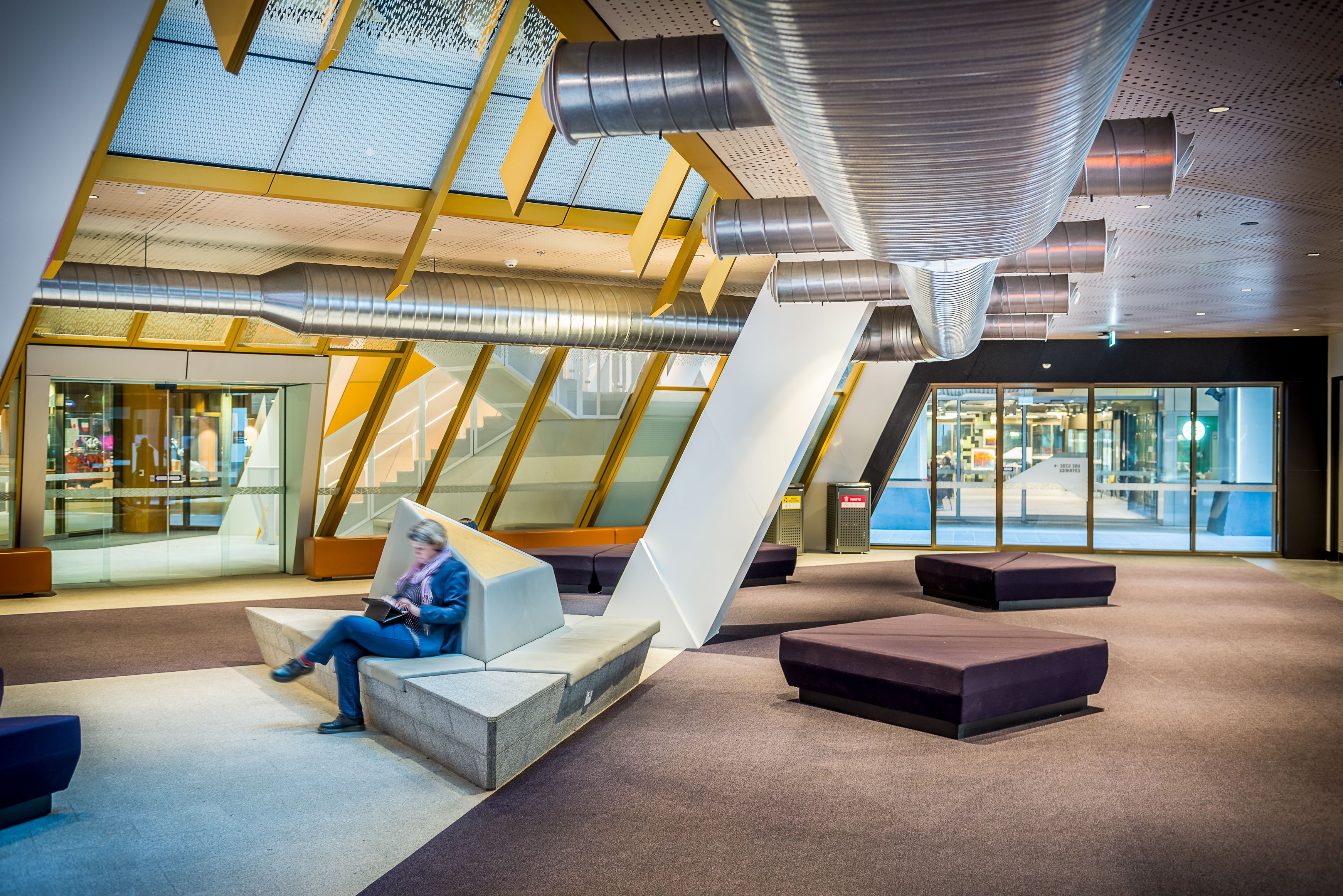 Modern lobby with exposed ventilation ducts, angled white supports, benches, and a seated person reading.