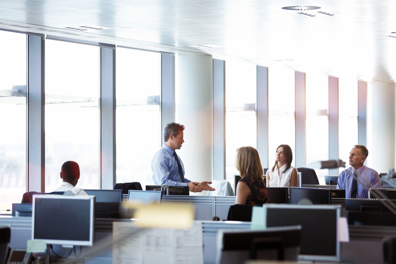 A group of colleagues in an office, sitting at desks while one colleague is standing and talking to them.