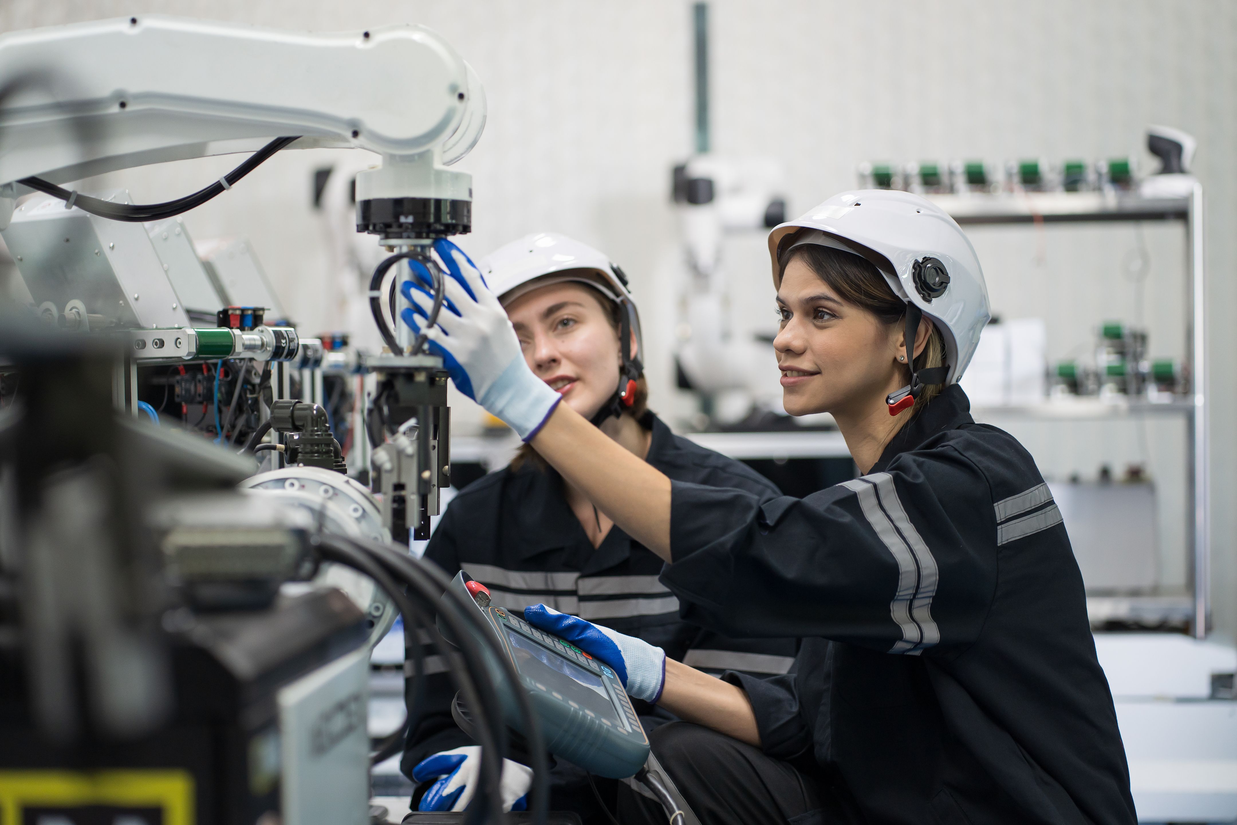 Two industrial workers wearing safety helmets and gloves inspect a robotic arm in a manufacturing facility.