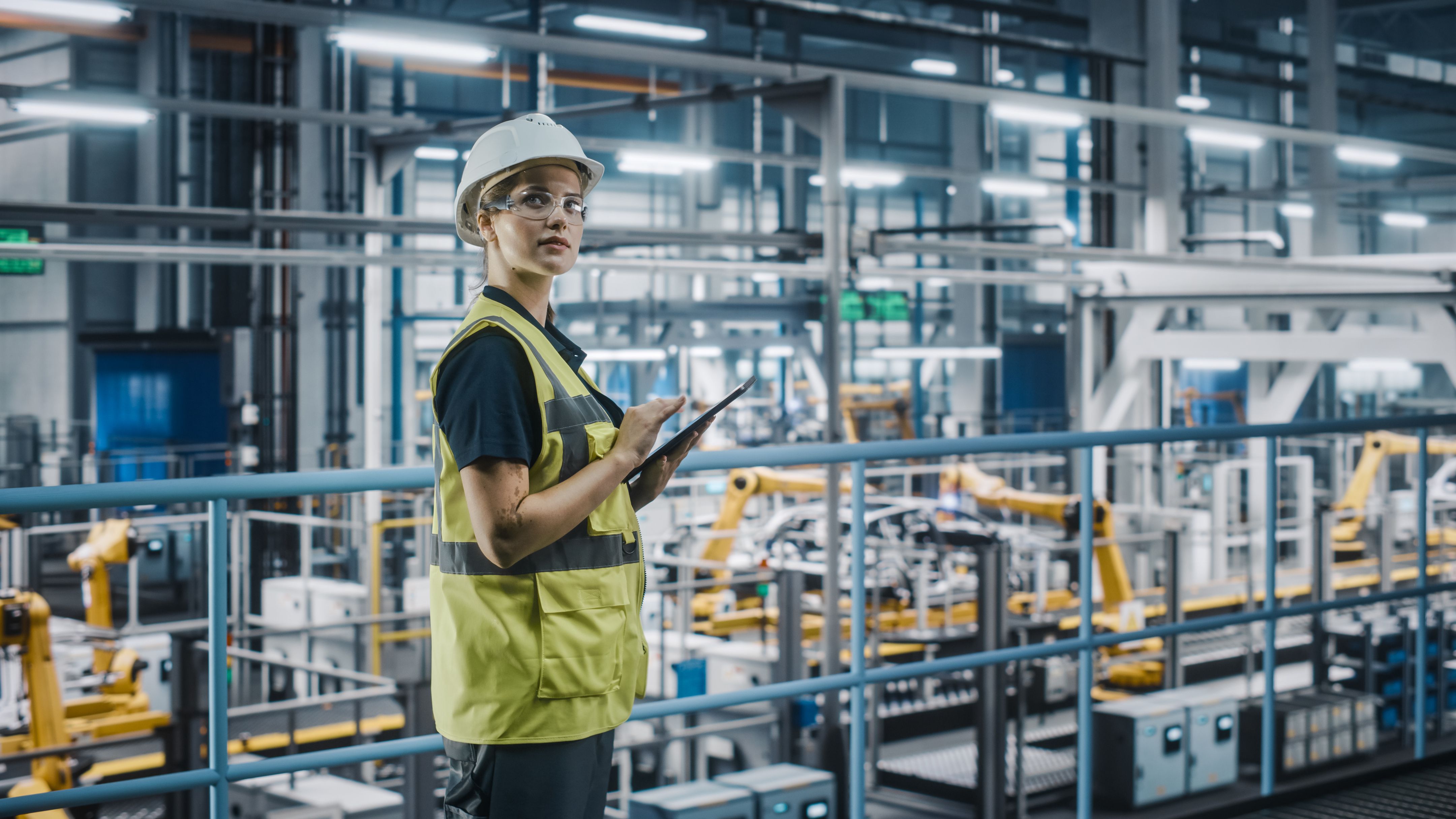 Female engineer with high visibility vest on an elevated walkway using a tablet to monitor production and workplace safety.