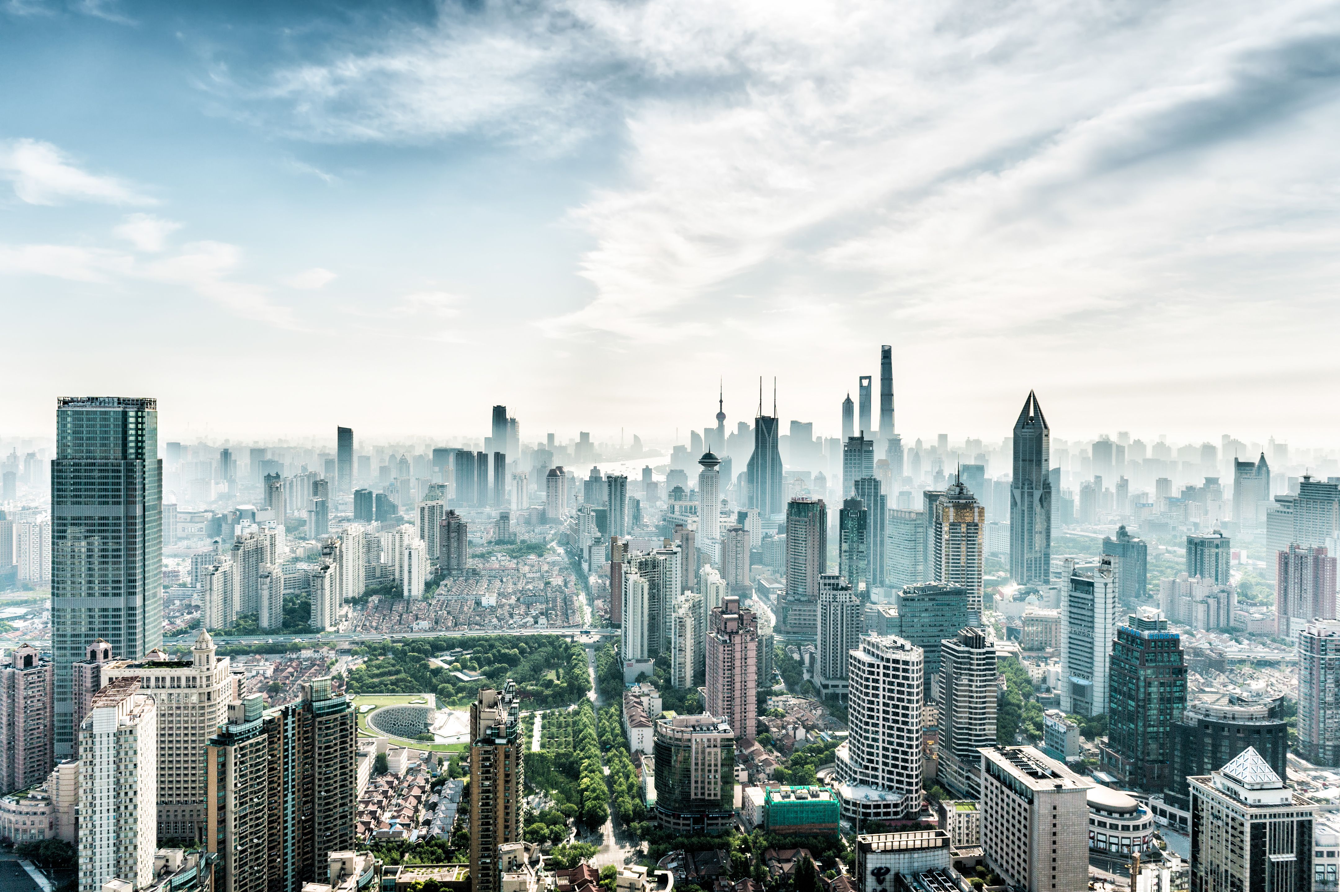 High-angle view of a sprawling modern city skyline with numerous skyscrapers under a bright, hazy sky.