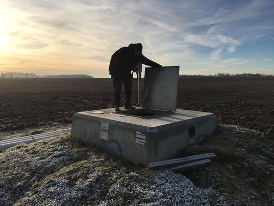Worker inspecting an access hatch on a concrete water well structure in an open field at sunrise.