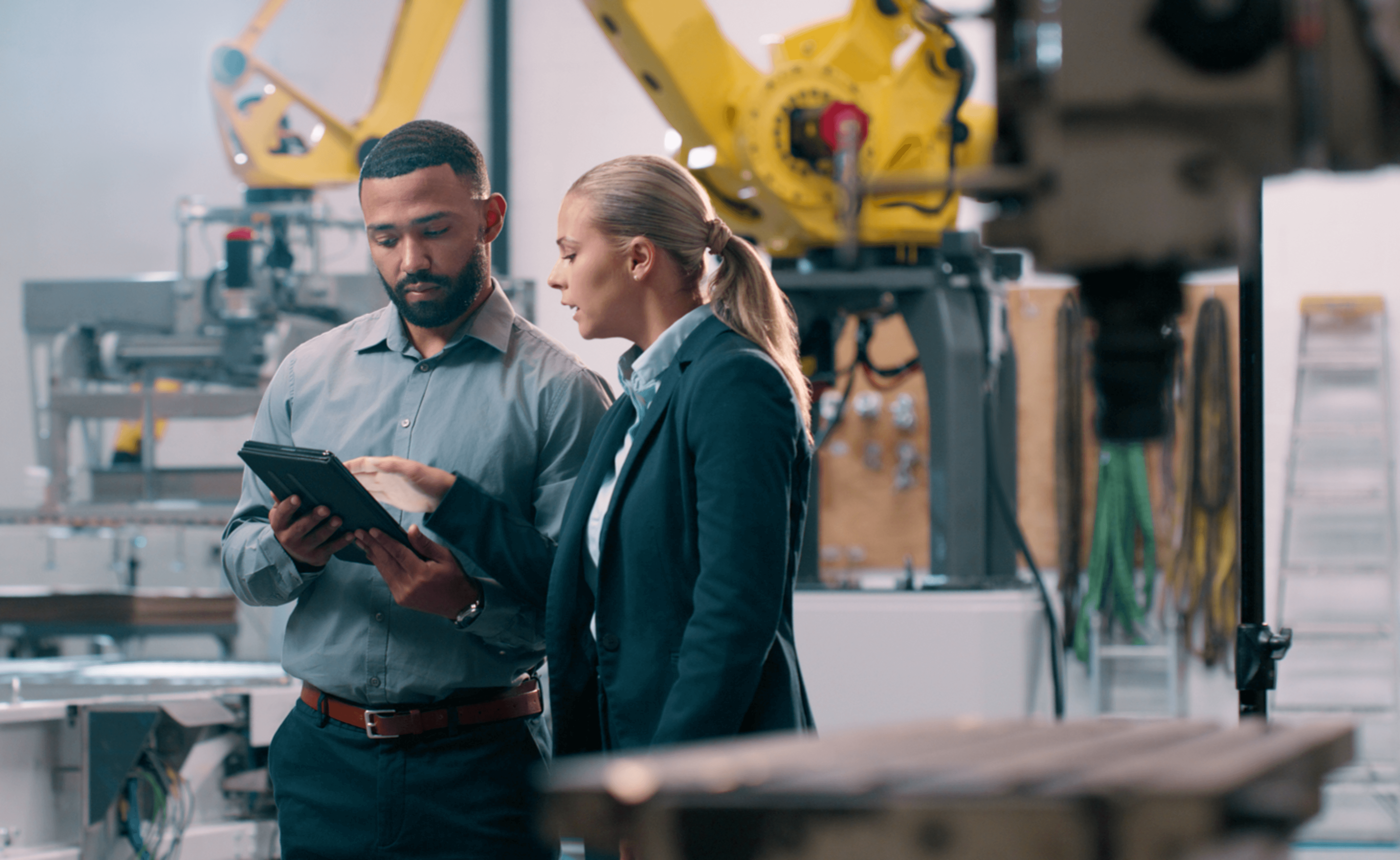 Two professionals review a tablet in a modern factory with yellow robotic arms in the background.