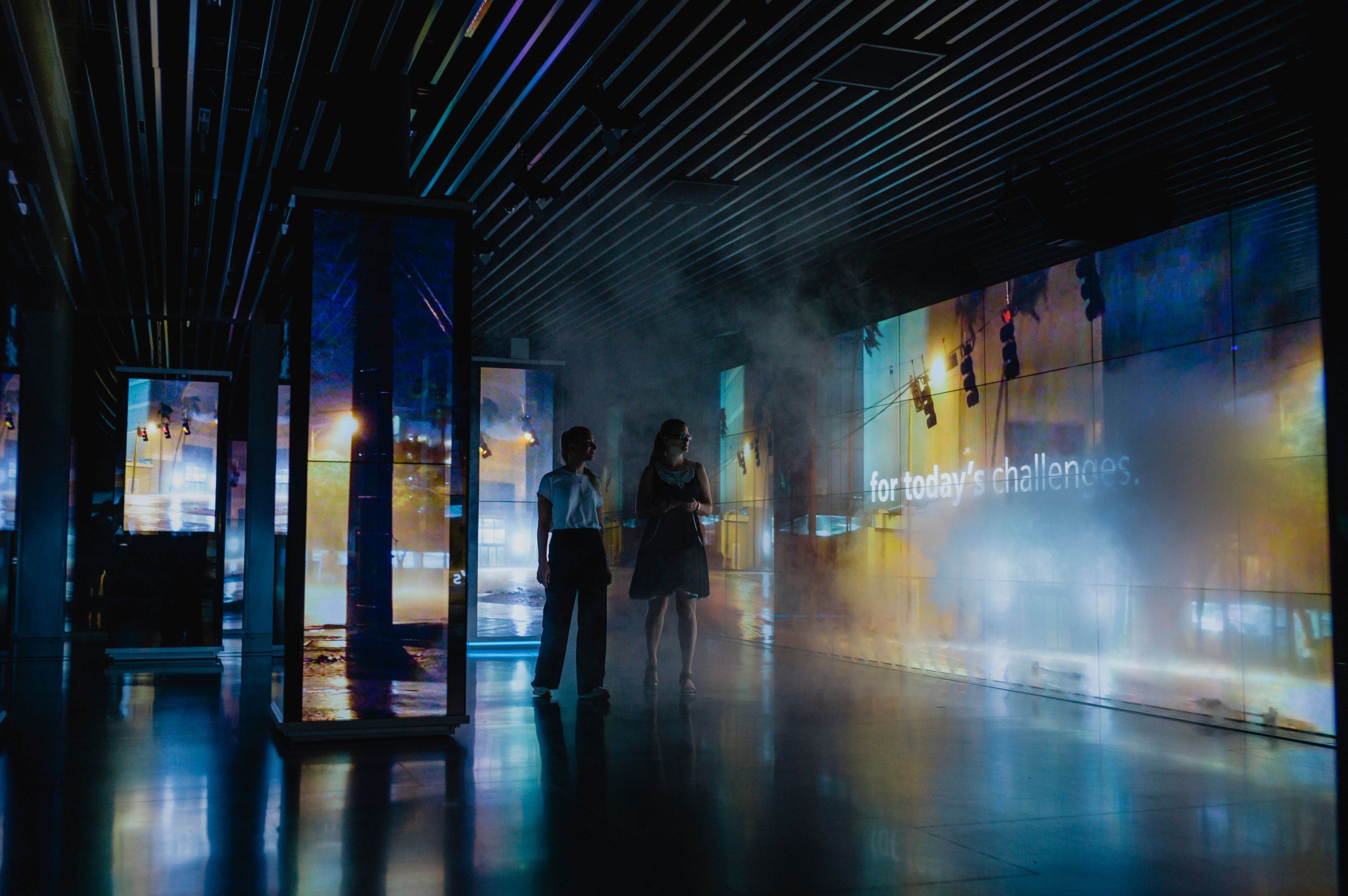 Two people stand in a dark immersive gallery with illuminated screens, mist, and the text “for today’s challenges.”