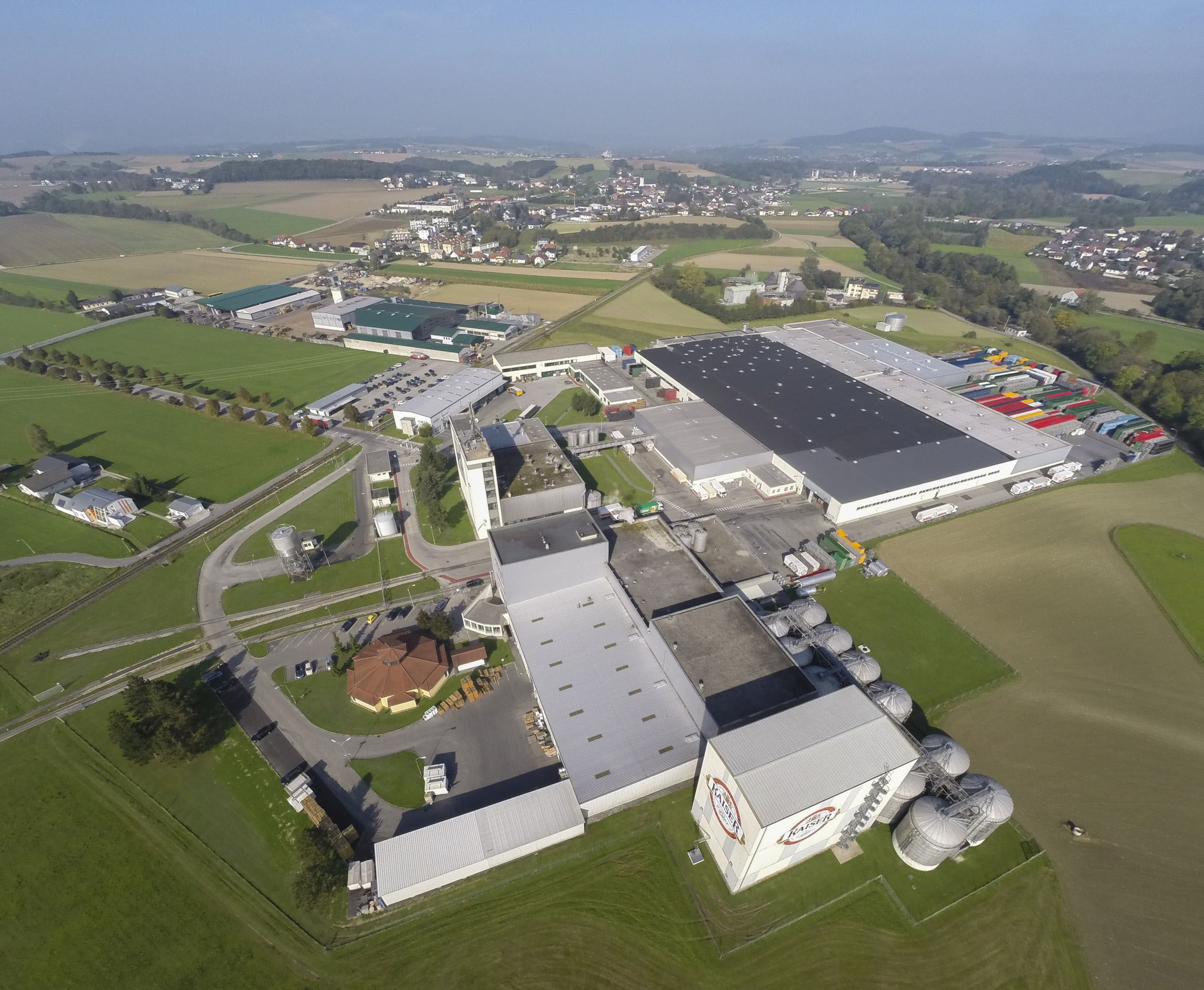 Luftfoto av Brau Union bryggerikompleks med produksjonsbygninger og solcellepaneltak omgitt av jordbruksland.