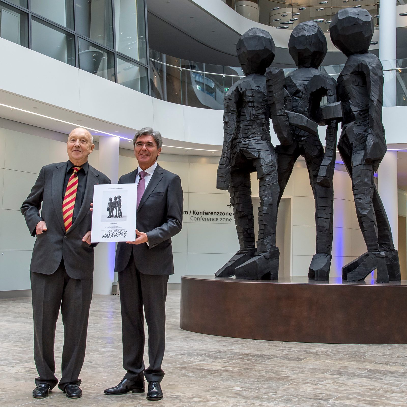Two people in suits hold a framed print beside four large faceted human sculptures in a modern atrium.