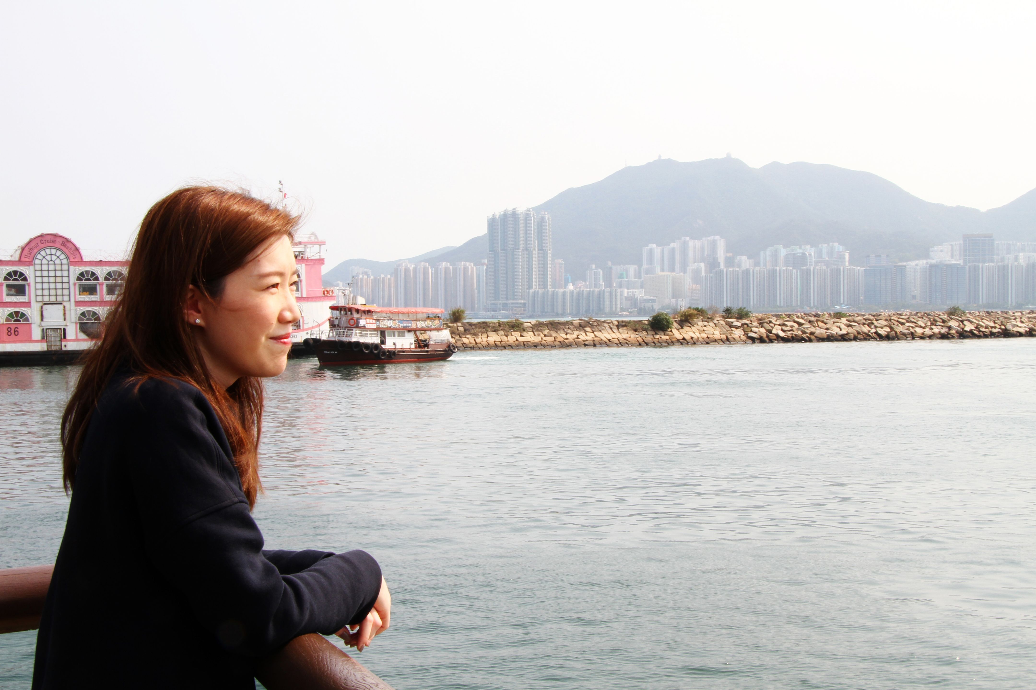 A woman stands on a bay in front of a city skyline, looking over the water