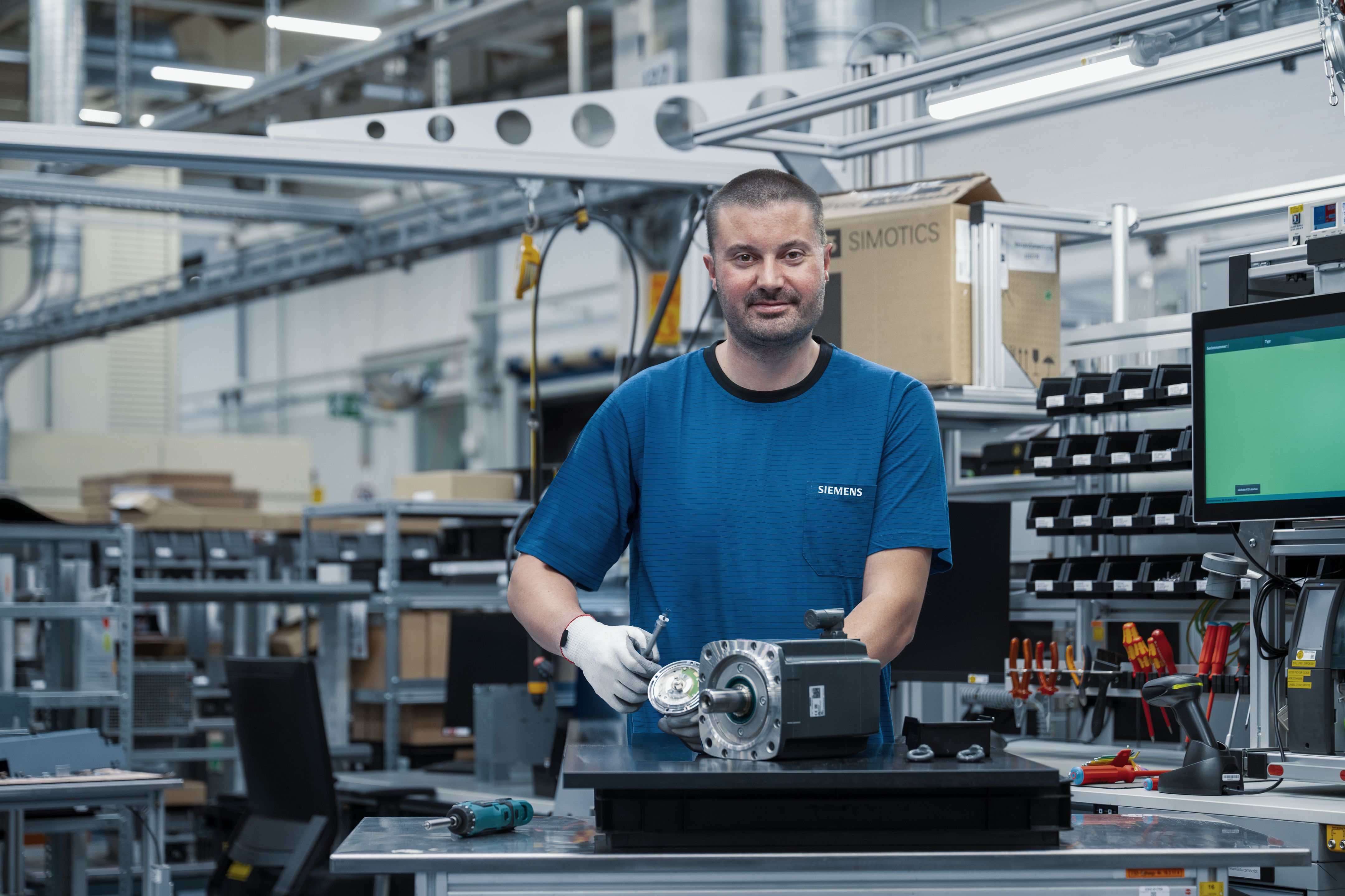 Person ved et arbejdsbord i et industrianlæg med en SIMOTICS-motor og komponent, hylder i baggrunden.