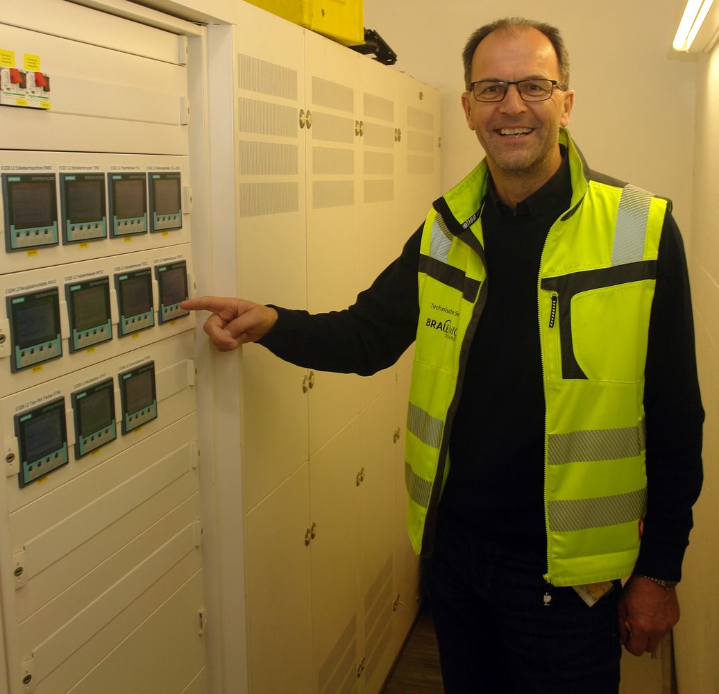 En mann i en høy synlighetsvest som peker på energimålere montert i et kontrollskap.