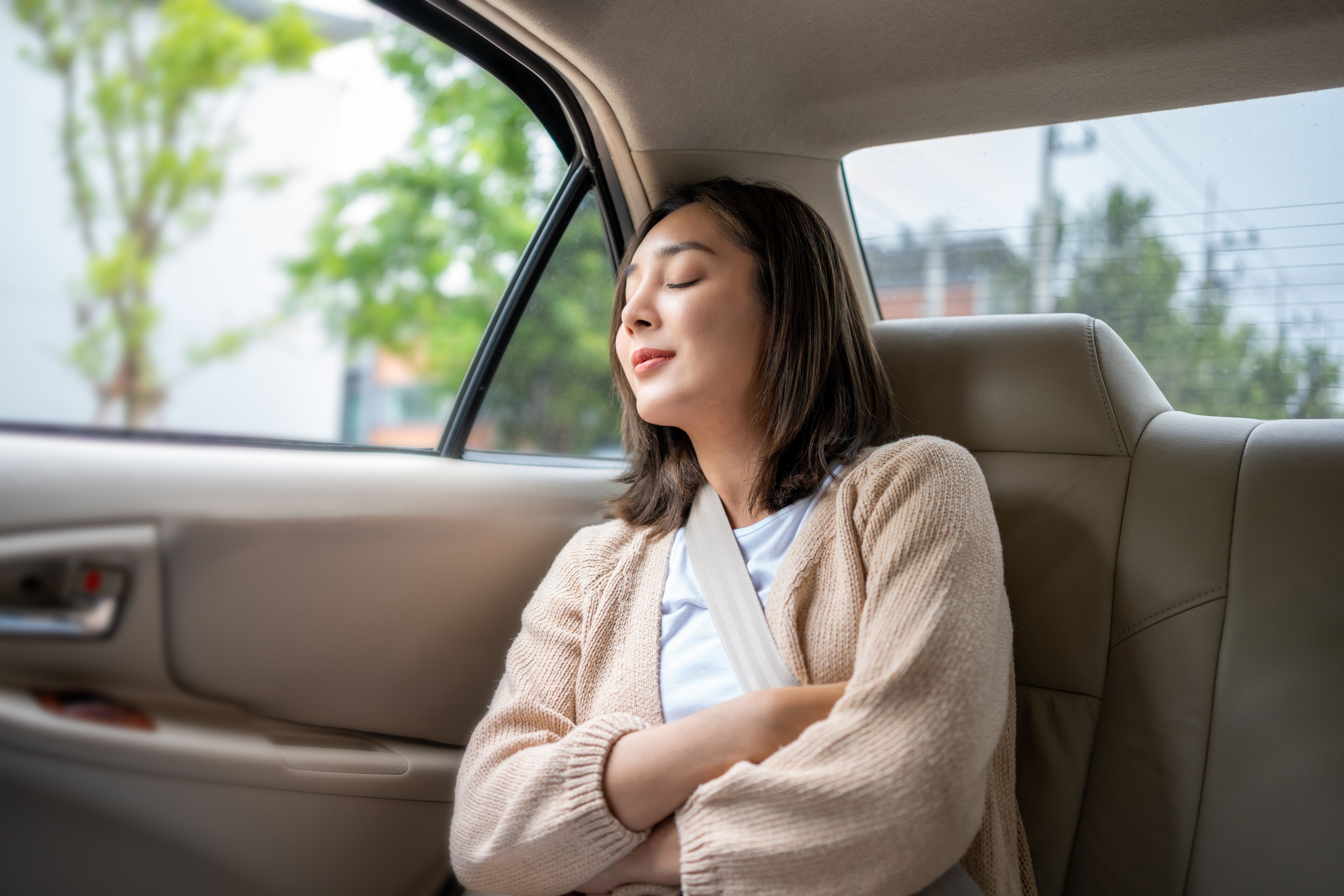 A young woman with her eyes closed, wearing a seatbelt and a light-colored cardigan, rests in the back seat of a car.