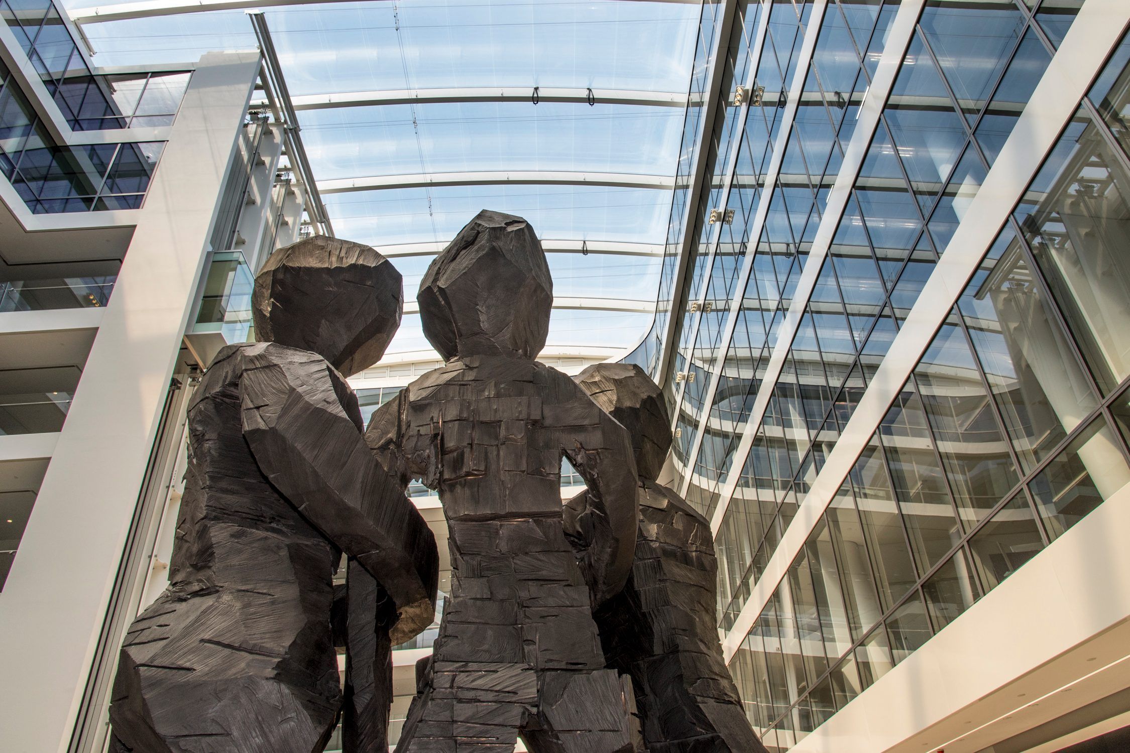 Four faceted human-like sculptures stand together beneath a glass-roofed atrium surrounded by office windows.