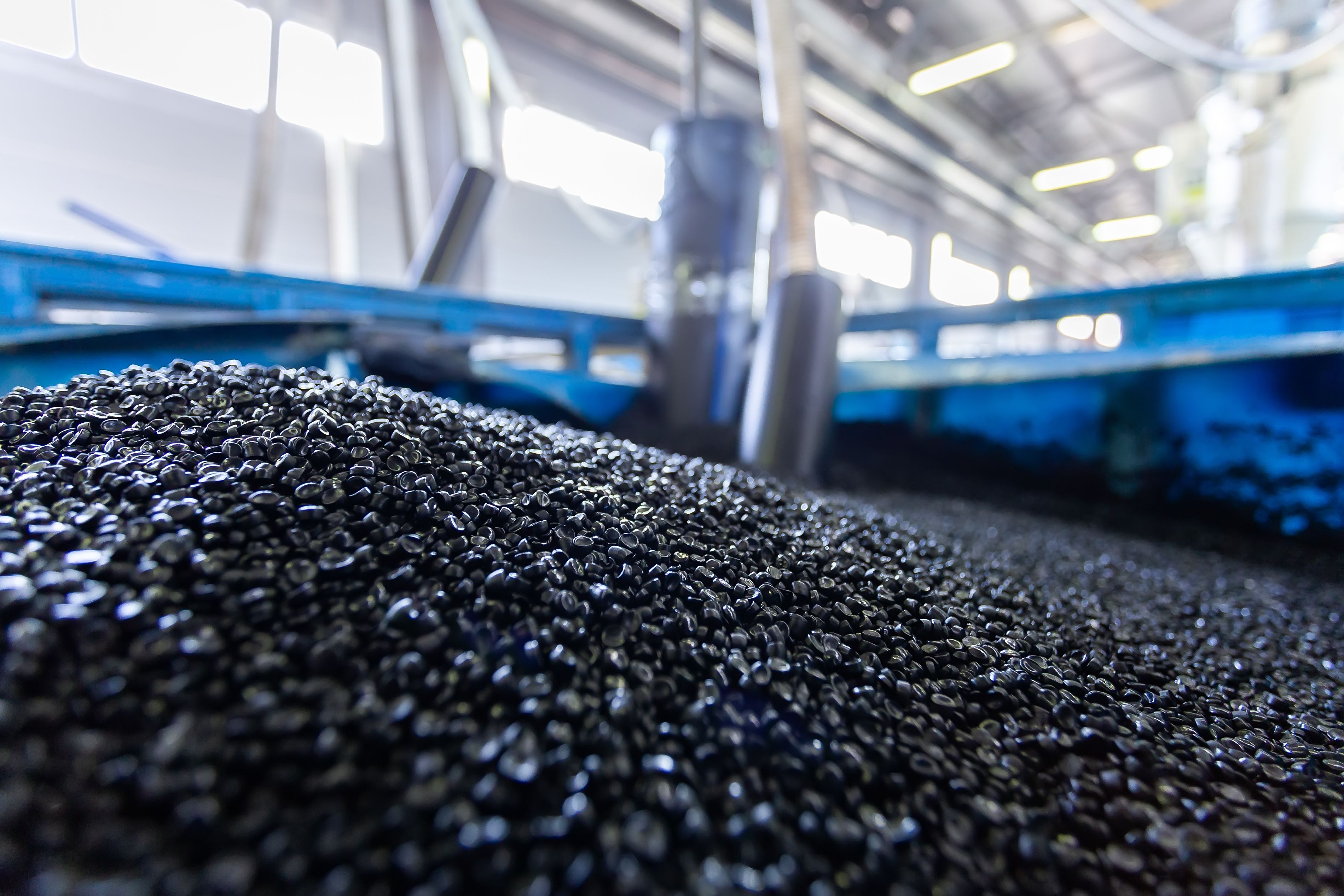 Close-up of black plastic pellets piled on a conveyor in an industrial facility with blurred machinery behind.