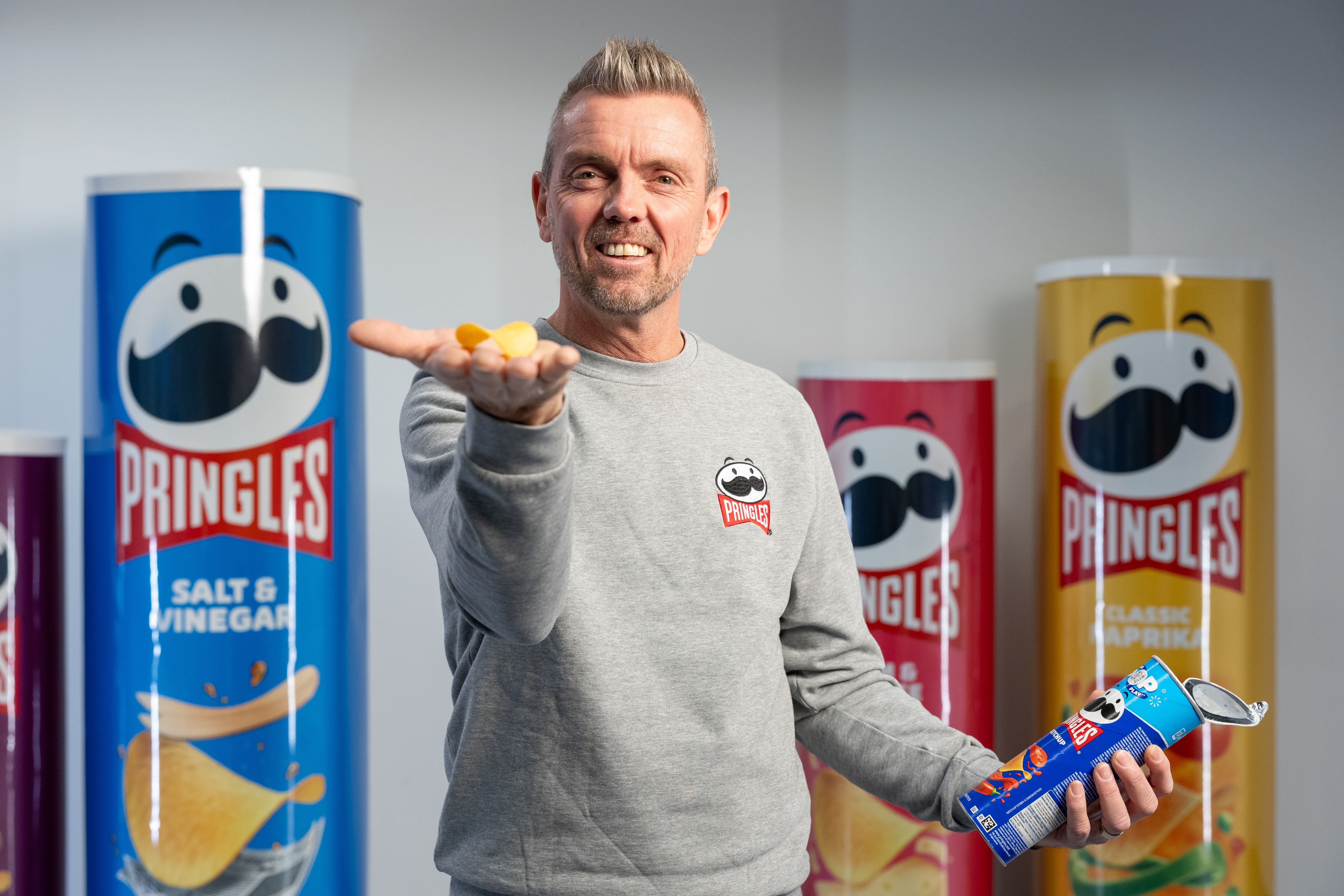En man ler och håller ut en handfull Pringles-chips i ena handen, stående framför överdimensionerade Pringles-behållare.