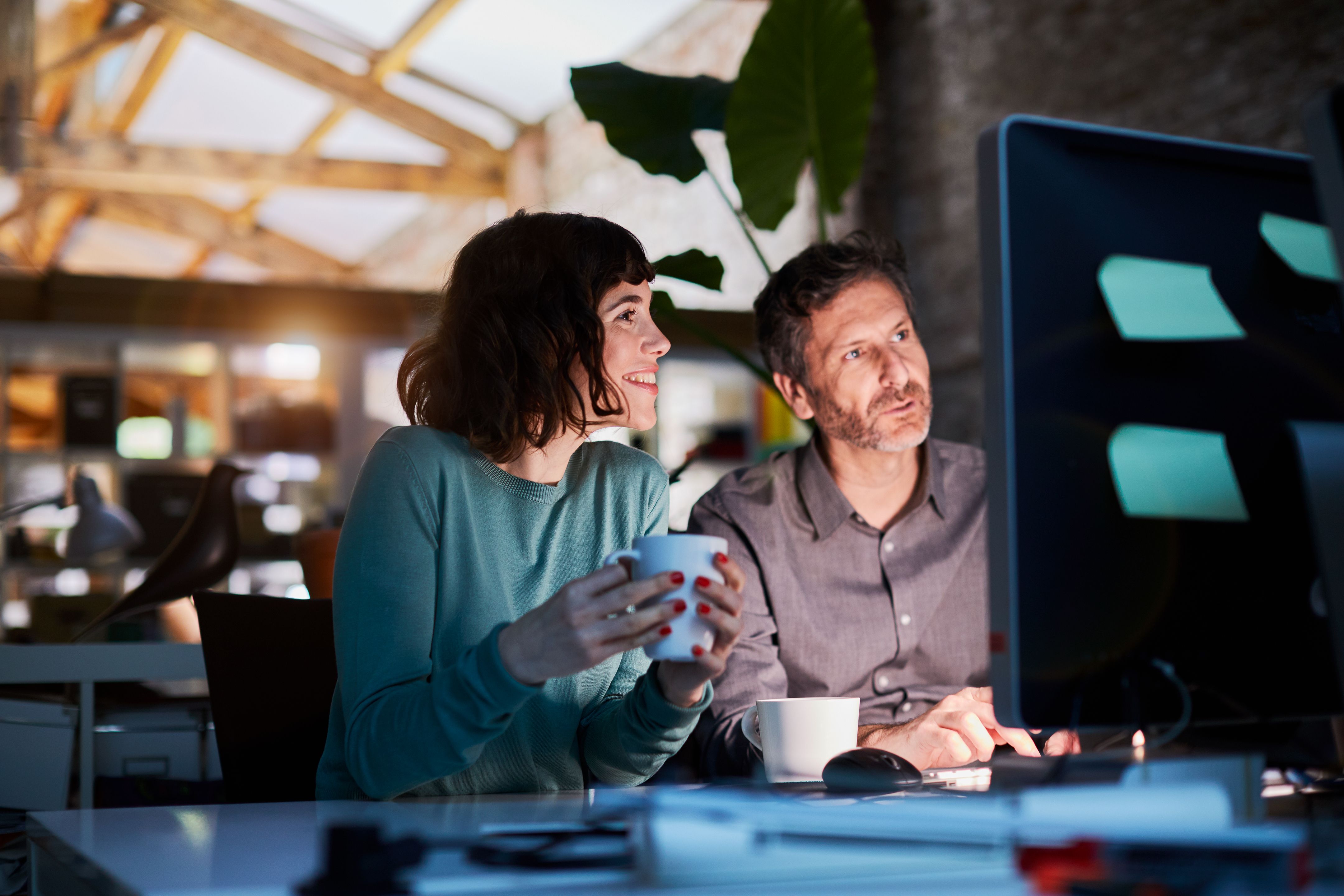 Two coworkers looking at a computer screen while drinking coffee, possibly tea. 