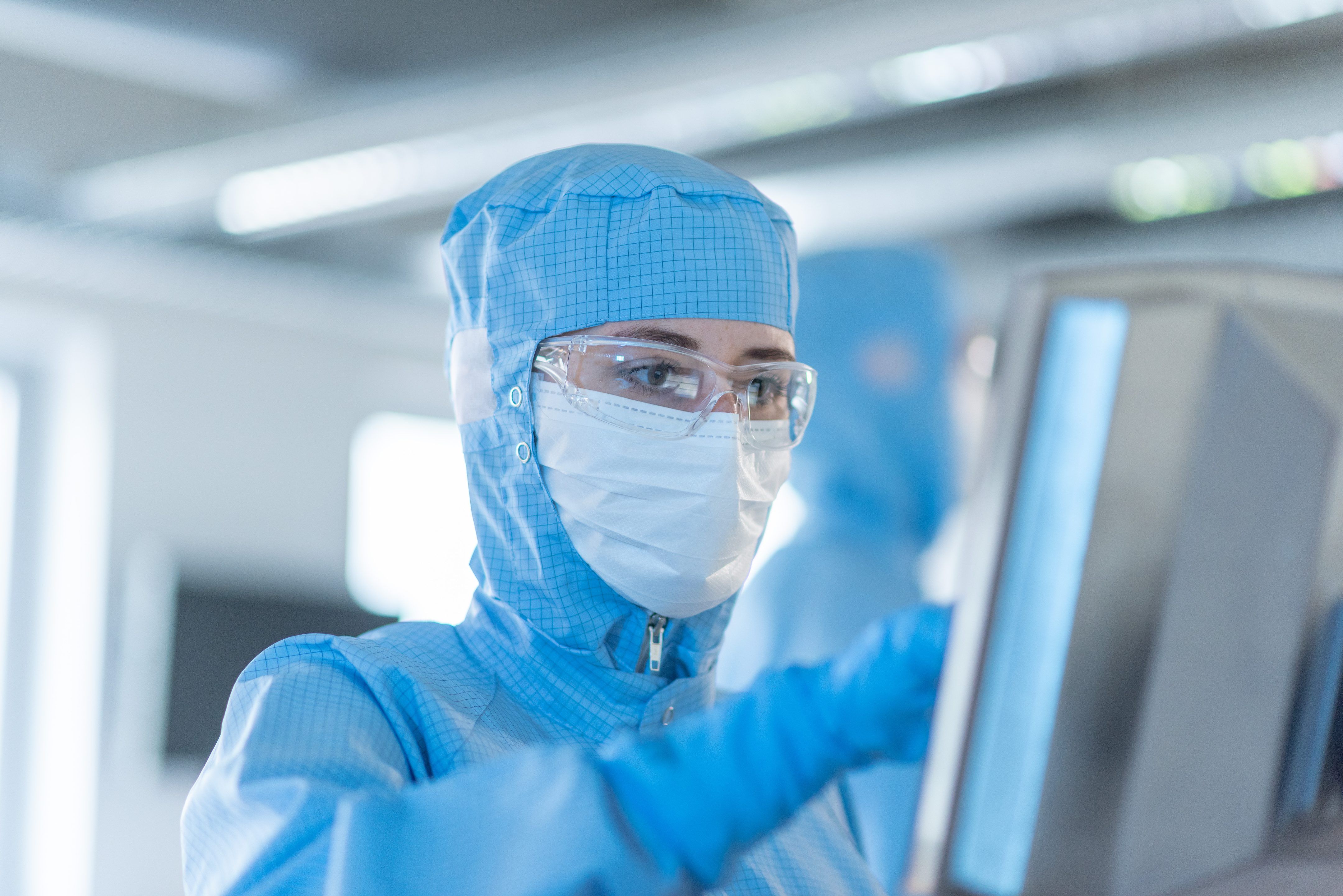 Worker wearing a cleanroom suit, mask, and safety glasses using a monitor.