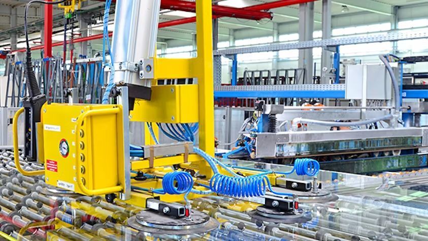 A tapadókorongokkal ellátott sárga robotkar egy gyárban lévő szállítószalagon lévő üveglapokat kezeli.