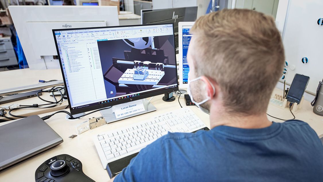 The picture shows an employee of the Siemens Electric Motor Factory in Bad Neustadt sitting at his desk. There are two monitors on the desk. On the left one you can see the digital twin of a motor, the second monitor is covered by the employee's head.