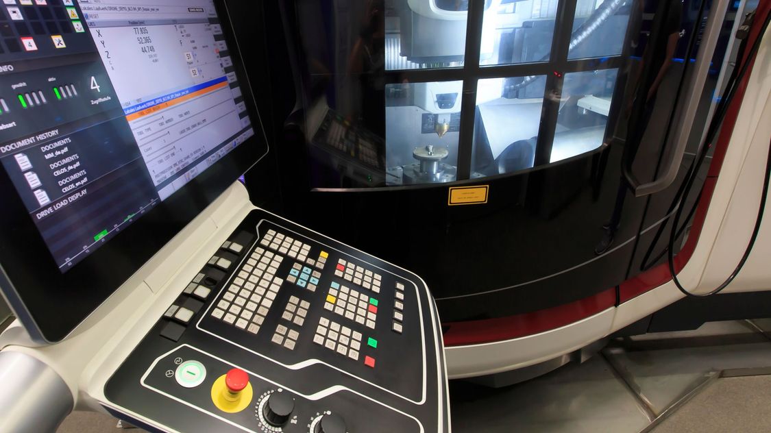 Foto einer Sinumerik-basierten Maschinensteuertafel mit einer Werkzeugmaschine im Hintergrund, in deren Arbeitsraum ein DED-Werkzeug für das Laserauftragsschweißen gerüstet ist.