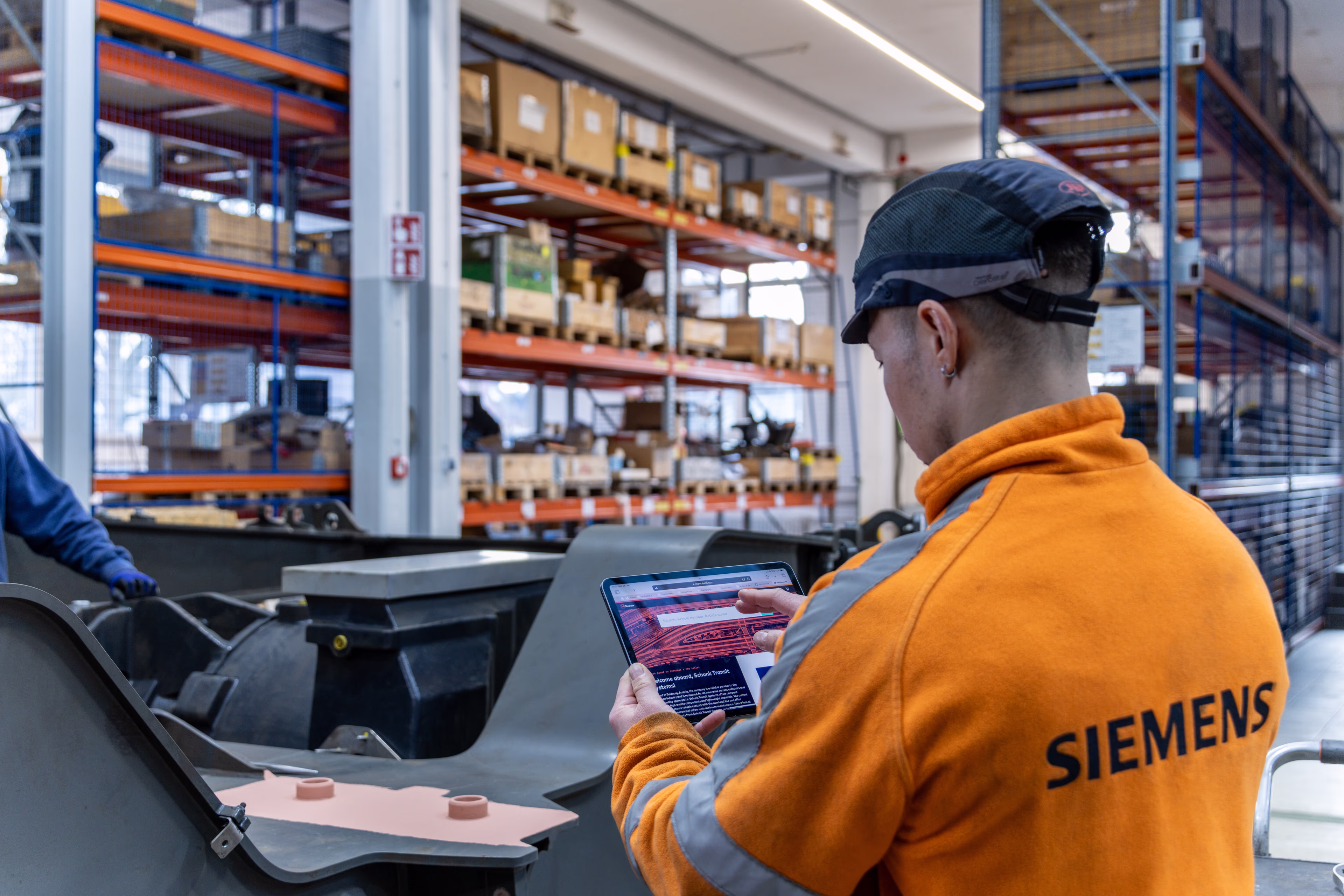 Ein Mitarbeiter, der eine Warnschutzjacke mit der Aufschrift „Siemens“ auf dem Rücken trägt, schaut auf ein Tablet. Im Hintergrund sind Regale zu sehen, und vor ihm liegen Bauteile. Siemens Mobility Bahnservices Instandhaltung