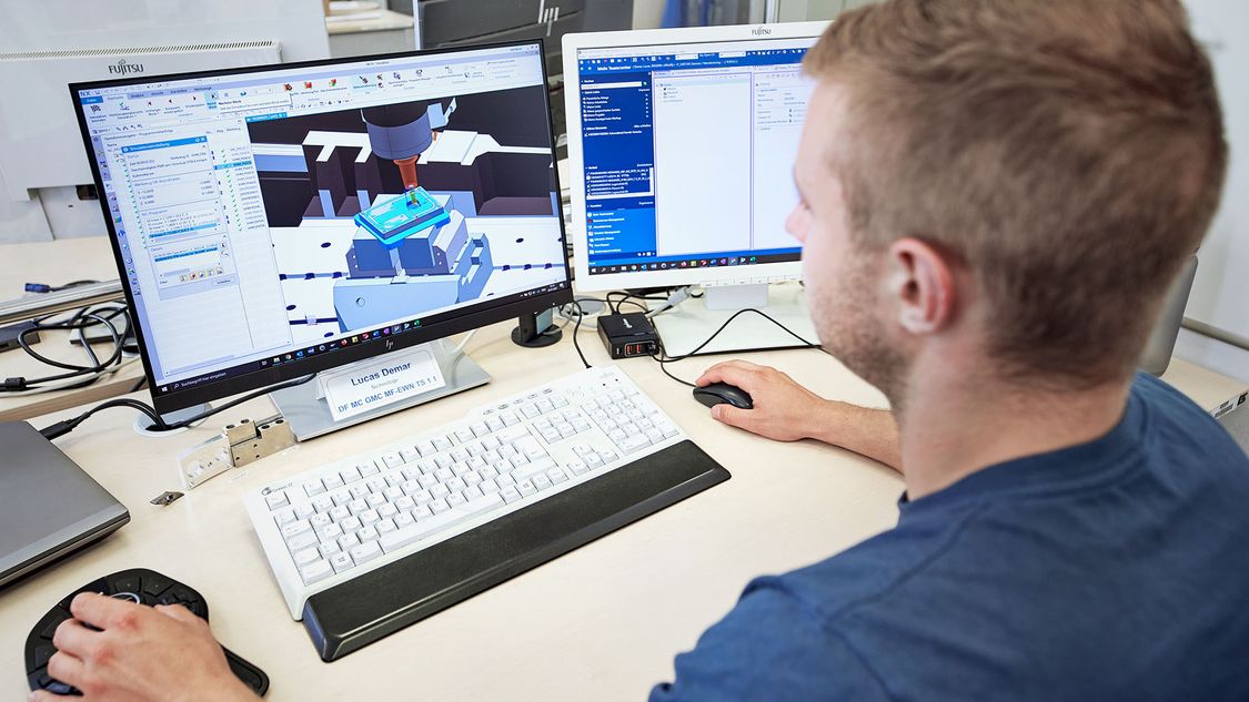 The picture shows an employee of the Siemens Electric Motor Factory in Bad Neustadt in front of a laptop with a second monitor connected. The display of the laptop shows the digital twin of a motor, the second monitor shows tables.