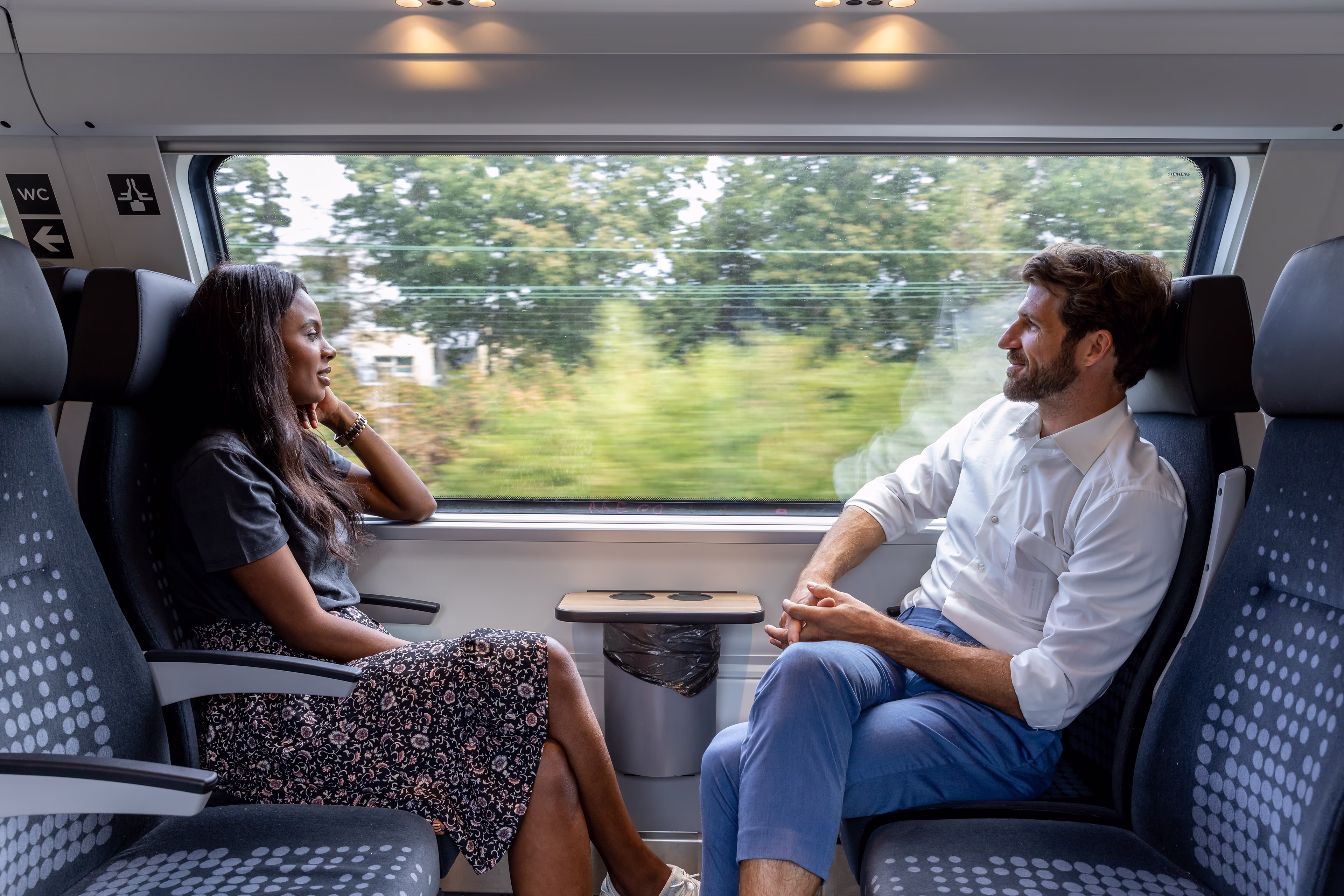 Zwei Fahrgäste sitzen in einem Zugabteil nebeneinander und schauen aus dem Fenster. Im Fenster sind digitalisierte Icons zu sehen. siemens mobility intelligent cctv keyvisual