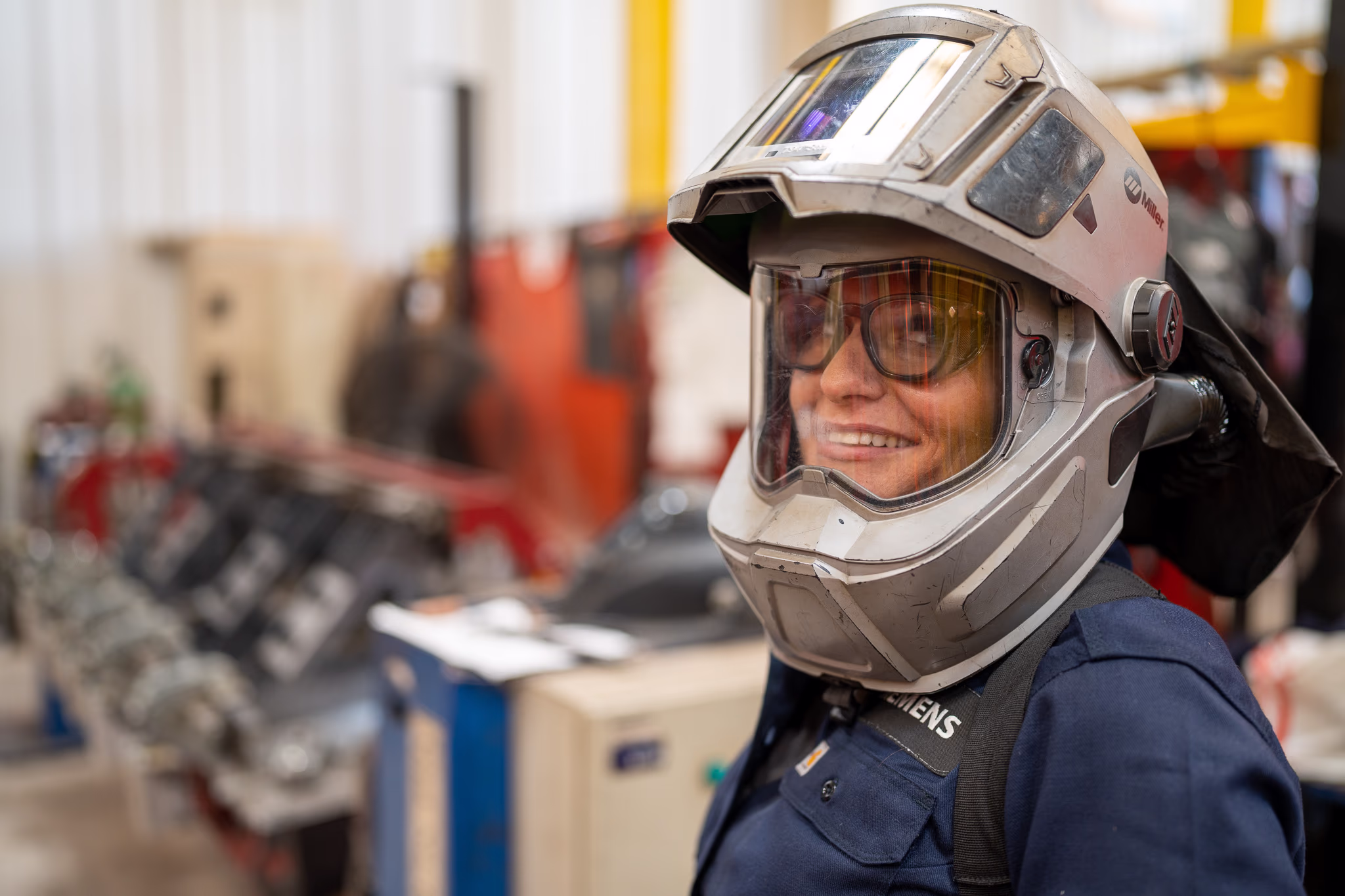 A smiling woman wearing a welding helmet and safety glasses looks to the right in a workshop environment, with "Siemens" visible on her work shirt. SMO-SAC-3