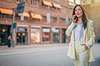 Eine Frau Mitte 30 in einem weißen Anzug steht neben einer Straße mit urbanem Charakter. Sie telefoniert mit ihrem Smartphone.