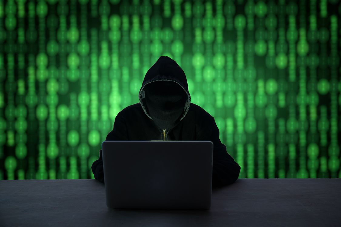Person completely concealed in dark clothing hacking on laptop with green matrix background