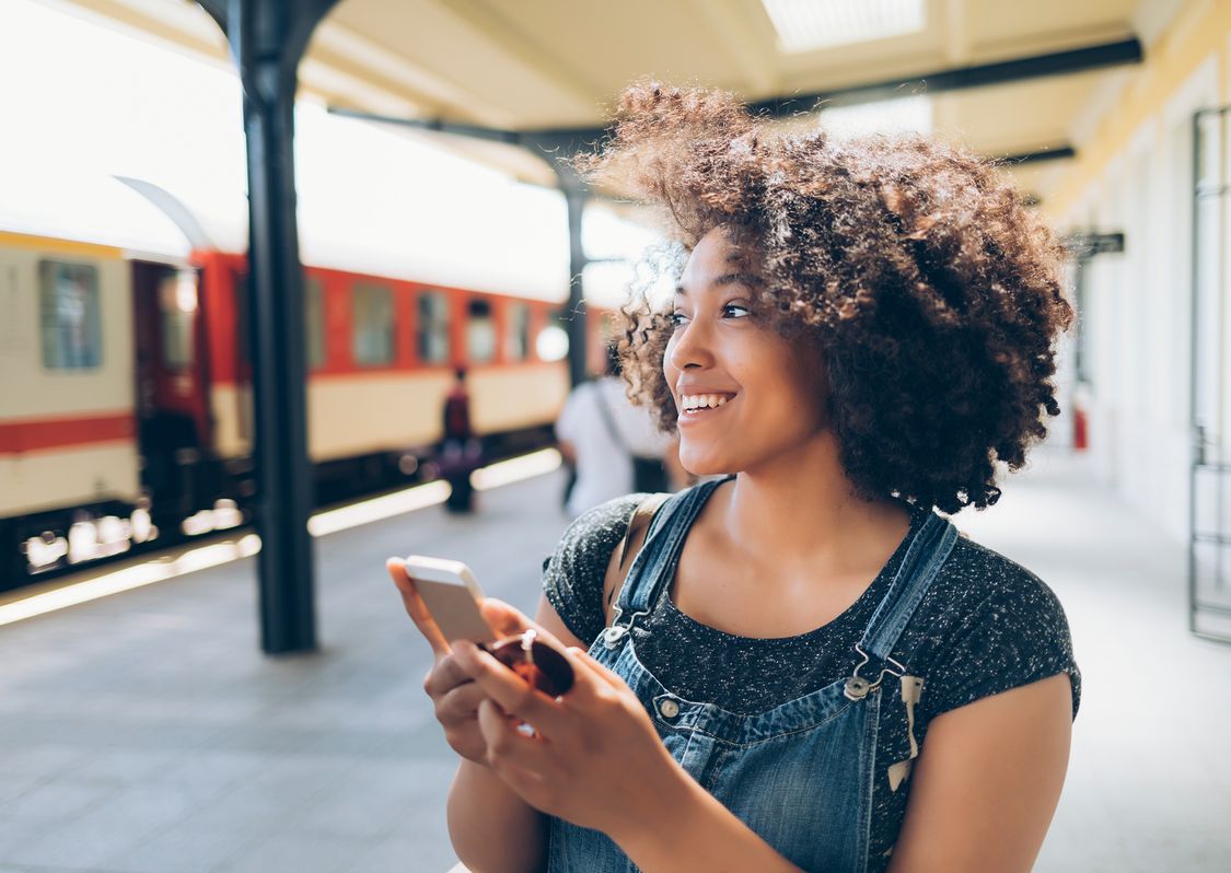 Woman curly hair is holding a smartphone an smiling while walking throu a railway station 