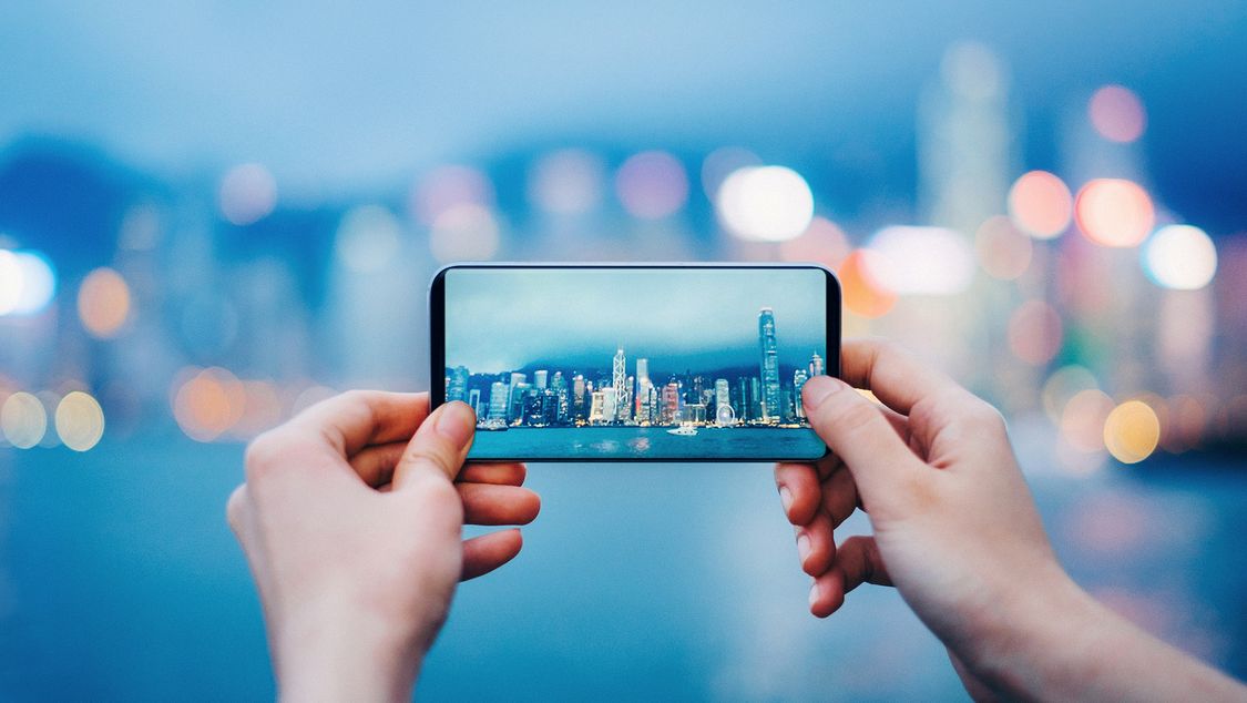 A pair of hands holds up a mobile phone against an out-of-focus skyline of a city. On the mobile device screen is the city in focus. 