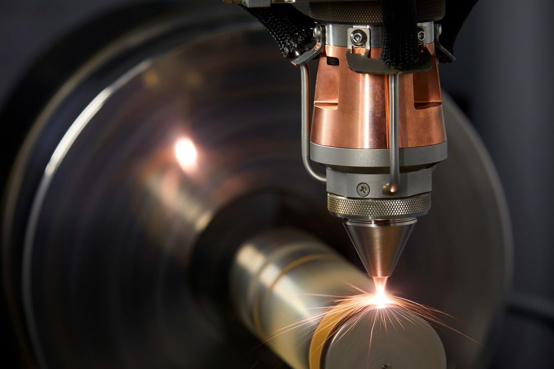 Photo: A round workpiece is clamped in the spindle of a machine tool, onto which additional material is applied by laser buildup welding with each revolution. 