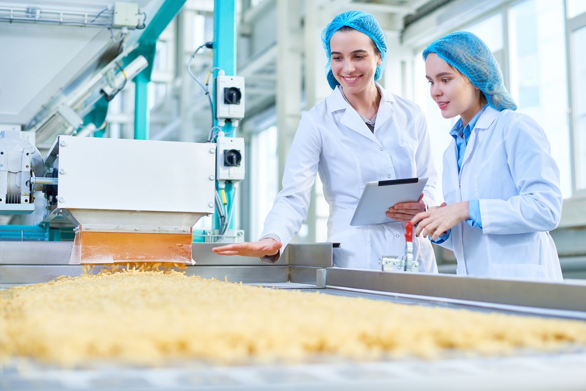 Portrait en plongée de deux jeunes femmes portant des blouses de laboratoire, debout près d'un convoyeur de macaronis dans un atelier de production propre