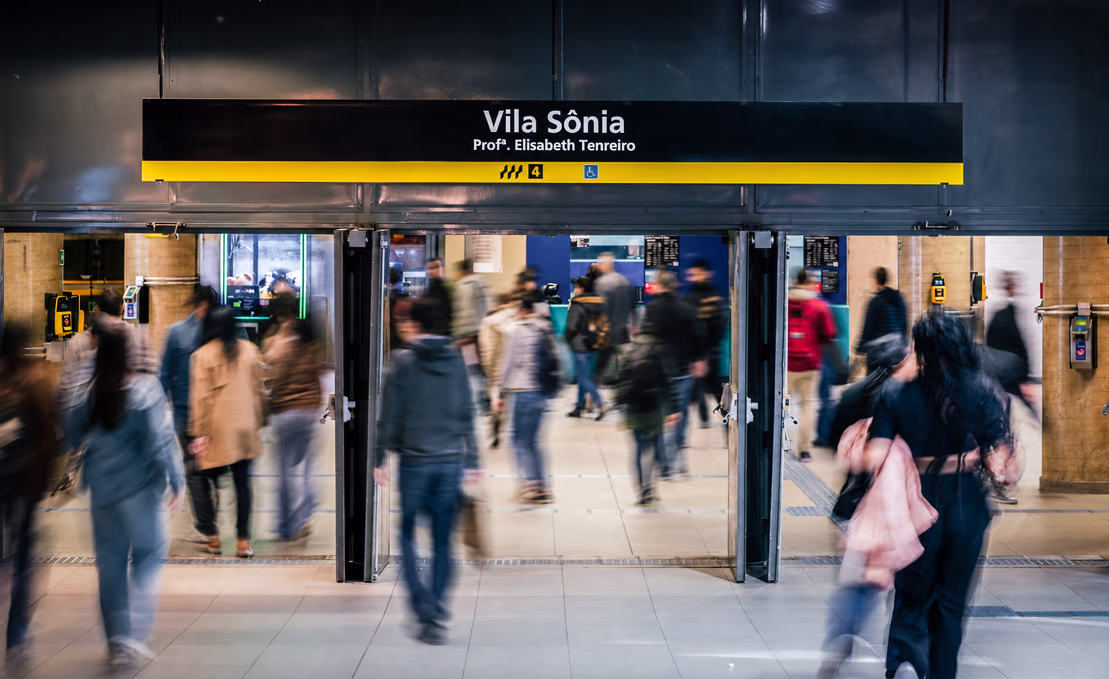 A bustling modern subway station entrance, "Vila Sônia Profª. Elisabeth Tenreiro," with blurred figures of people moving through automatic doors. sao-paulo-subway