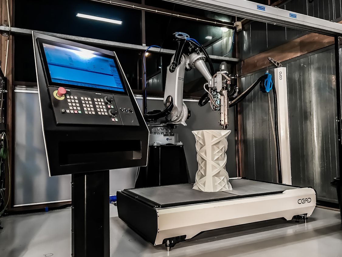 Foto eines Maschinentisches, auf dem Roboter ein stark strukturiertes, vasenförmiges Werkstück per Additive Manufacturing fertigt. Im Vordergrund eine Bedientafel mit SINUMERIK-CNC