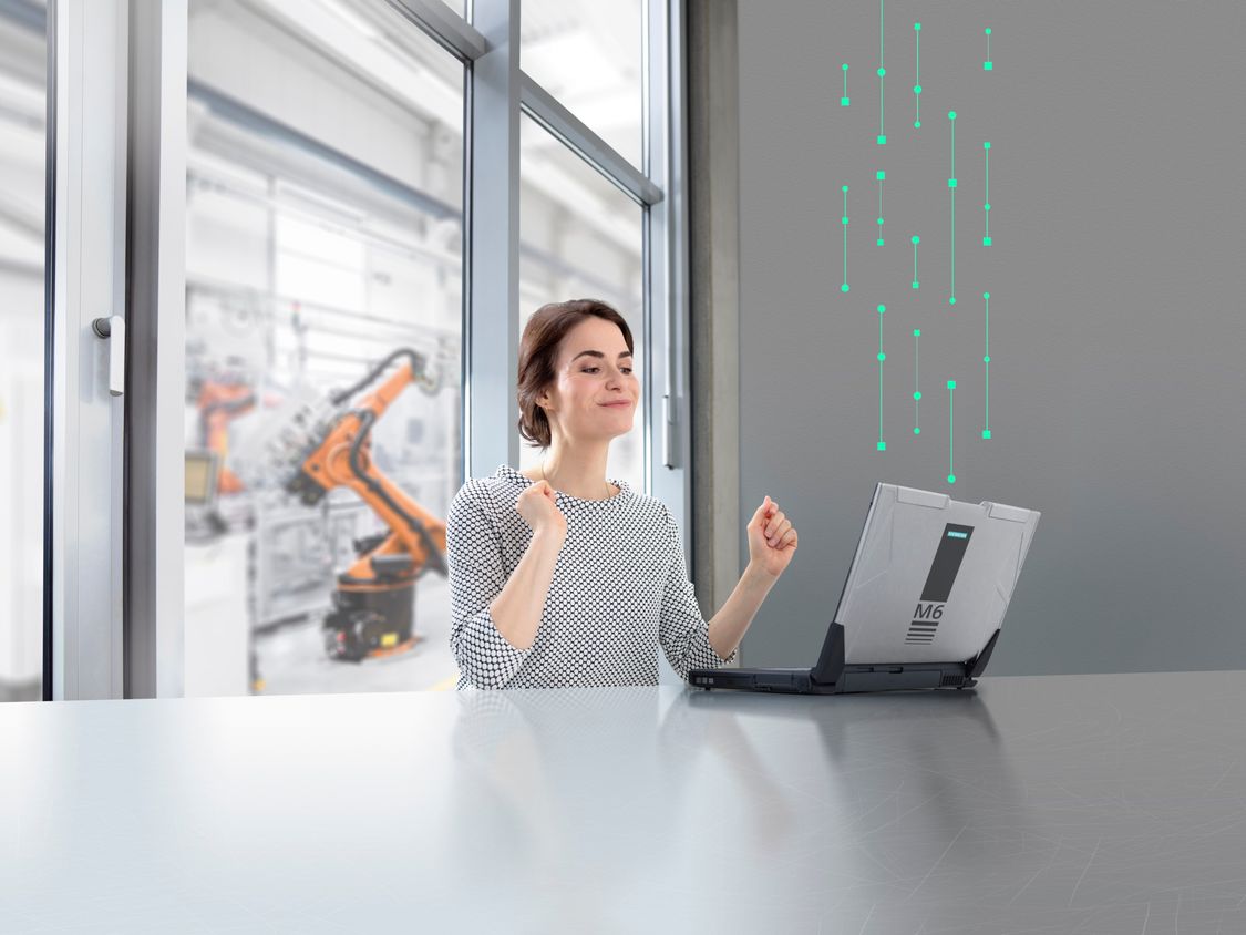 A young woman sits in front of her laptop in an office. Digital elements create a connection from the laptop to a production plant with robots in the background.