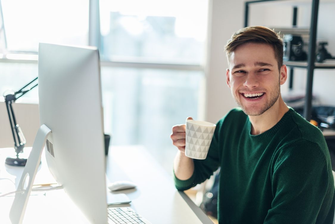 A student sits at a PC and smiles at the camera, holding a cup of coffee