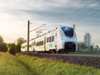 Mireo Smart drives under an overhead line through nature, with a high-rise building in the background. Siemens Mobility Mireo Smart
