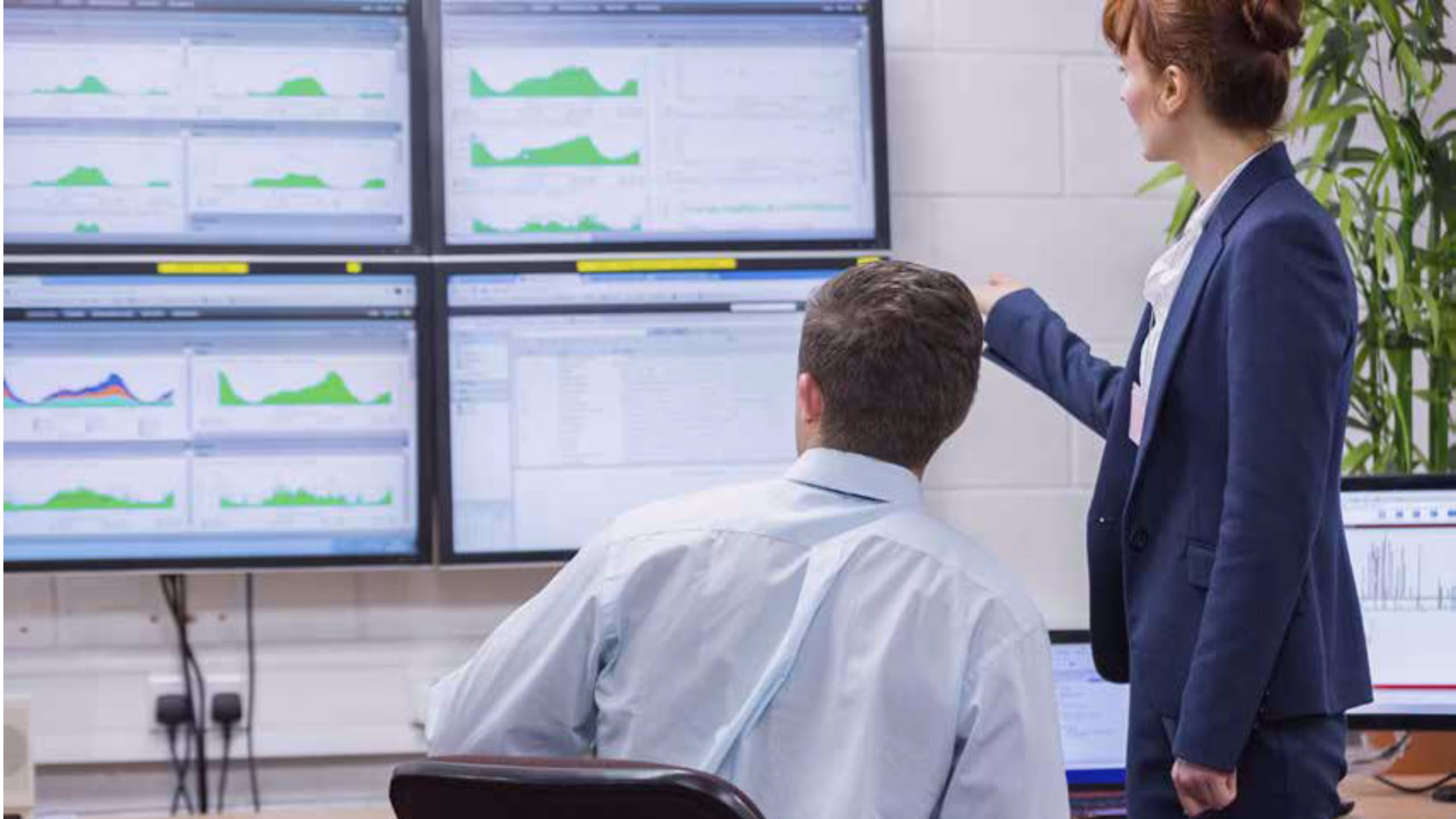 A man and a woman are looking at four monitors showing various graphs and statistics A man and a woman are looking at four monitors showing various graphs and statistics