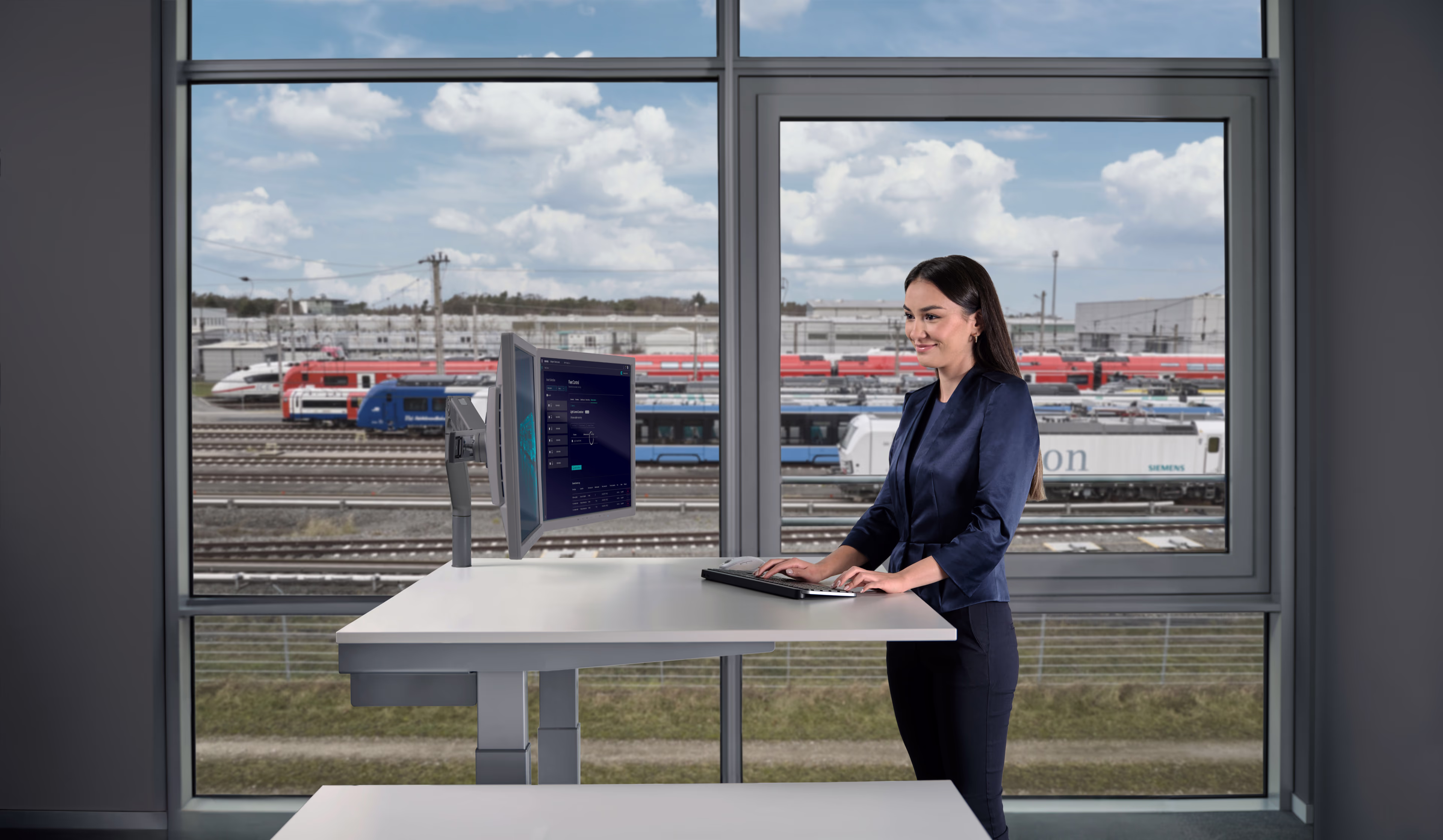 Eine Frau steht vor einem Fenster und arbeitet am Computer. Im Hintergrund sind verschiedene Schienenfahrzeuge von Siemens Mobility zu sehen. Siemens Mobility Digitaler Zugbetrieb Key Visual