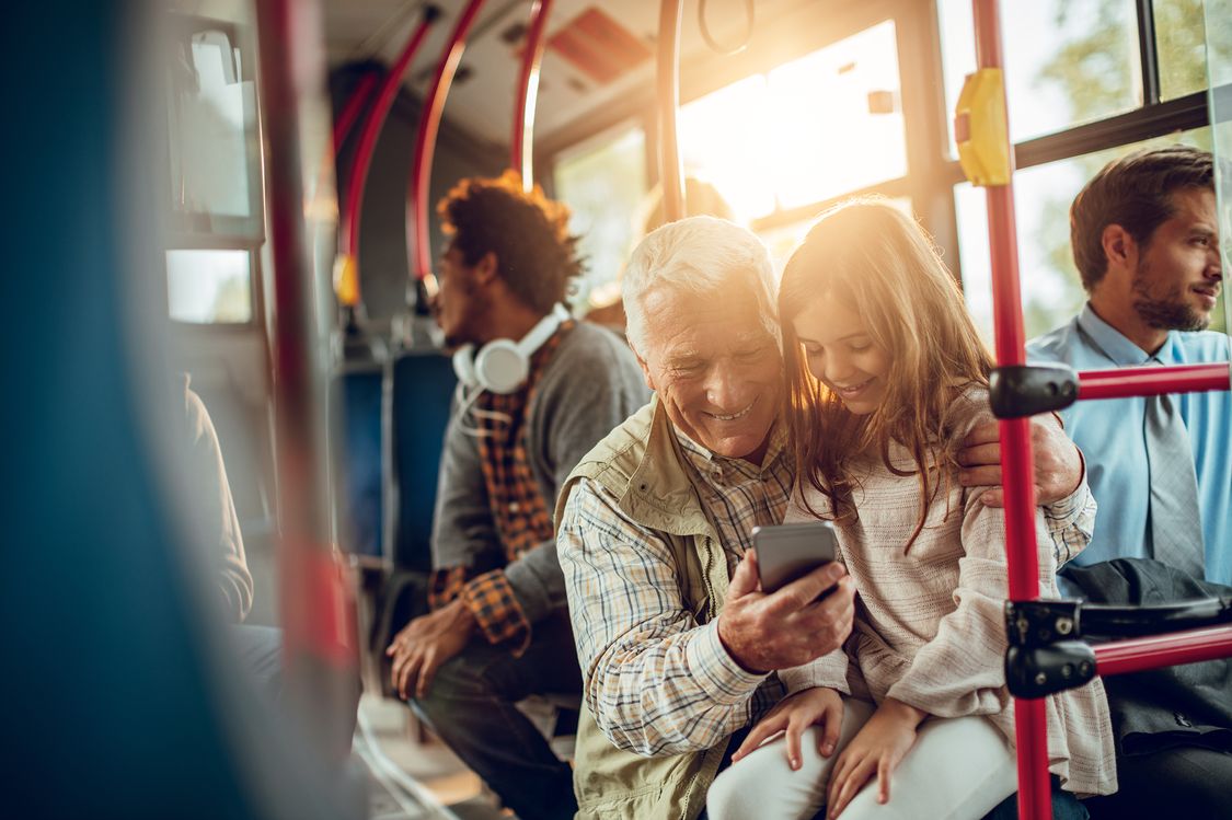 Grandfather and Granddaughter are sitting on a bus while smiling and looking at a smart phone
