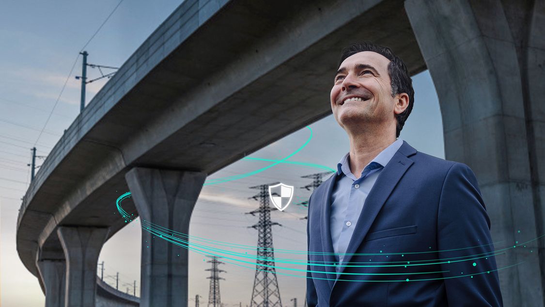A man stands in front of an elevated highway with electrical power lines in the background. The digital layer has four icons representing the different stages of implementing a robust cybersecurity regime (identify, protect, detect, manage) for critical infrastructure networks. 