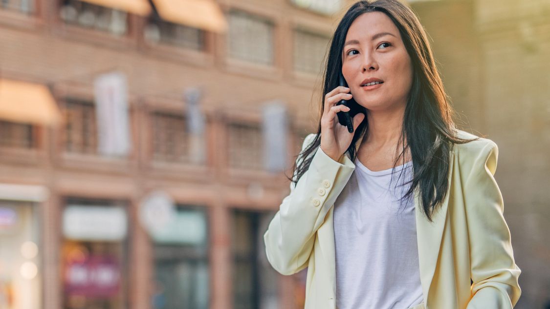 A woman in her mid-30s in a white suit stands next to a typical city street. She’s making a call on her Smartphone.