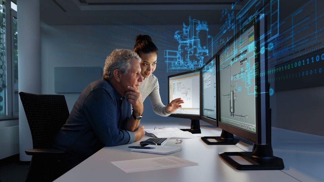 Two engineers look at plant data on multiple monitors, which is visible as a hologram in the background