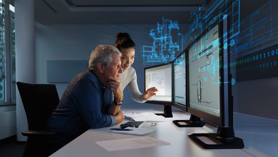 Two engineers look at plant data on multiple monitors, which is visible as a hologram in the background