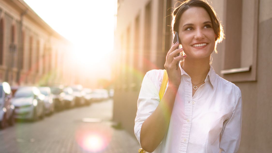 En ung kvinna står intill en gata som ser ut som en typisk stadsgata och ringer ett samtal på sin smartphone