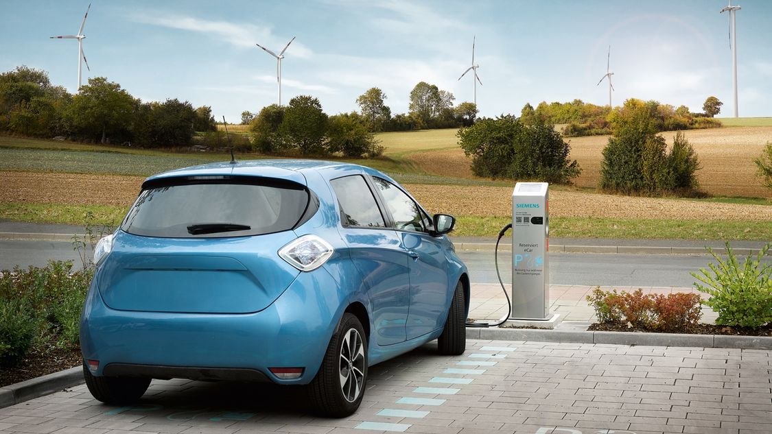 An electric car charges at a charging station, in the background there are wind turbines on a hill