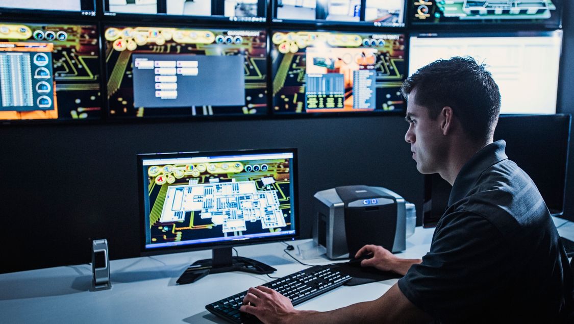 Operator in a control room working in front of a monitor on his desk, a monitor wall in the background.