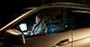 A man sits in a parked car at night, remotely working with a notebook that streams stylized data